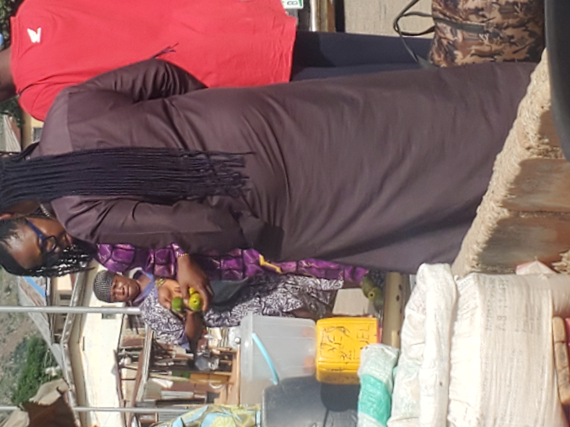 In Gwong, Nigeria, a woman with dark braided hair and glasses, wearing a dark brown robe, stands at an outdoor market. She is holding several small, green fruits in her hands. To her left, another woman in a patterned purple and white outfit is visible, also holding fruits. In the foreground, there are large white sacks, a yellow plastic stool, and a translucent bucket. Behind the women, buildings with metal roofs and a red shirted individual are visible. The ground appears to be paved.