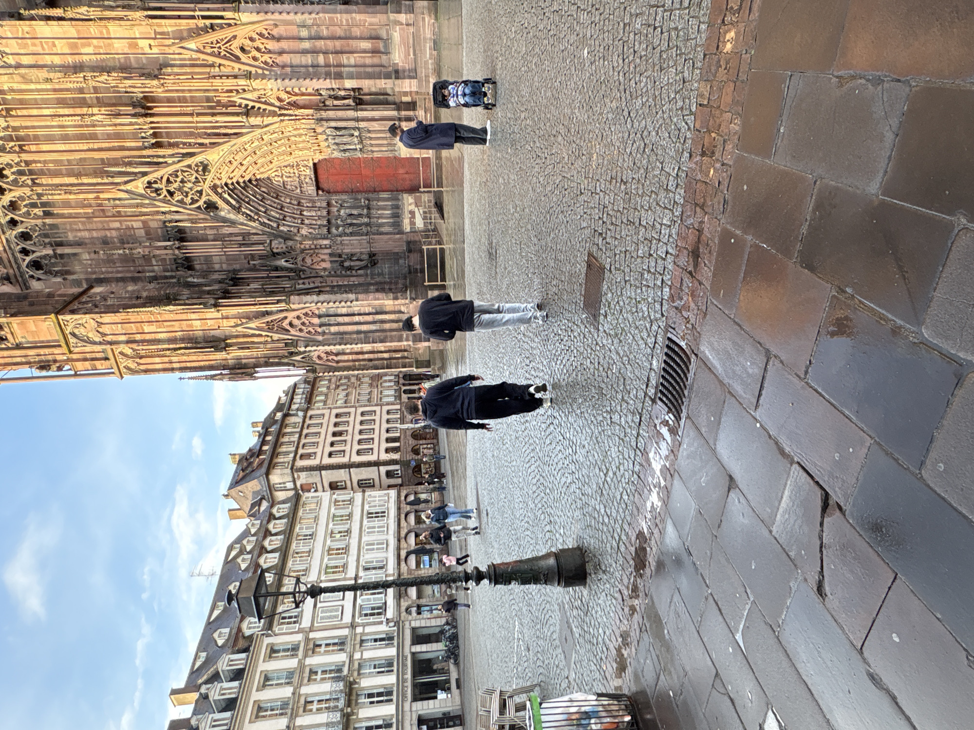 The image shows a view of the Place de la Cathédrale in Strasbourg, France. The Strasbourg Cathedral is visible in the upper left corner, and the surrounding historical buildings can be seen along the left side of the image. The ground is paved with cobblestones and larger, rectangular stones. Several people are present in the scene, some walking and one pushing a stroller. A lamppost stands in the lower left quadrant. A trash can with chairs is located in the lower left corner. The sky is visible in the upper left, with a few clouds.