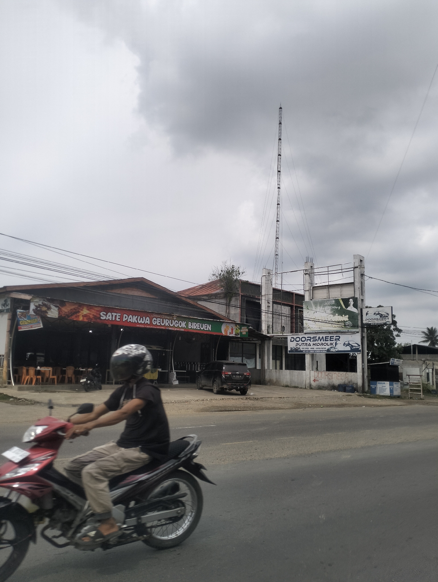 The image depicts a street scene under a cloudy sky. In the foreground, a person is riding a red motorcycle. Behind the motorcycle, a building with a large banner displaying "SATE PAKWA GEURUGOK BIREUEN" is visible. Further down the street, another building has signs that read "DOORSMER PUTRA HIDROLIK" and other text. A tall radio tower stands in the background. The environment appears to be a commercial area with various businesses and infrastructure.