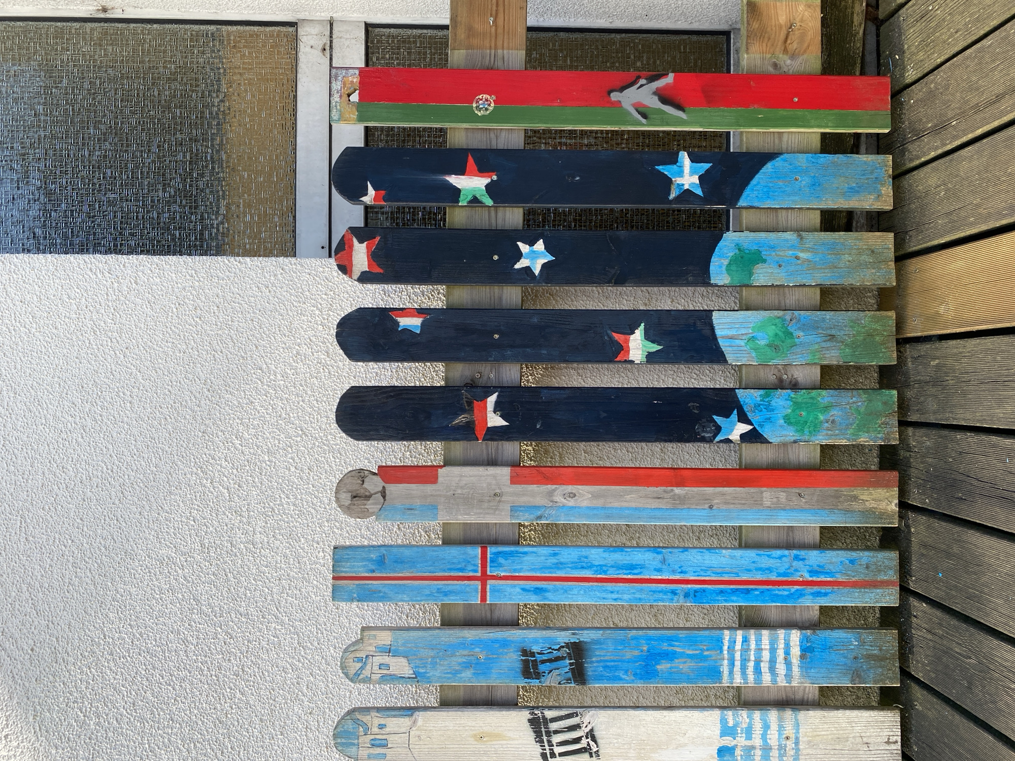 The image shows a row of painted wooden fence pickets in Altenkirchen, Germany. The pickets are decorated with various designs, including buildings, stripes, stars, and a silhouette of a person. The fence is positioned in front of a textured white wall and a window with a textured glass. The ground in front of the fence is made of wooden planks. The lighting suggests it is daytime with sunlight casting shadows.