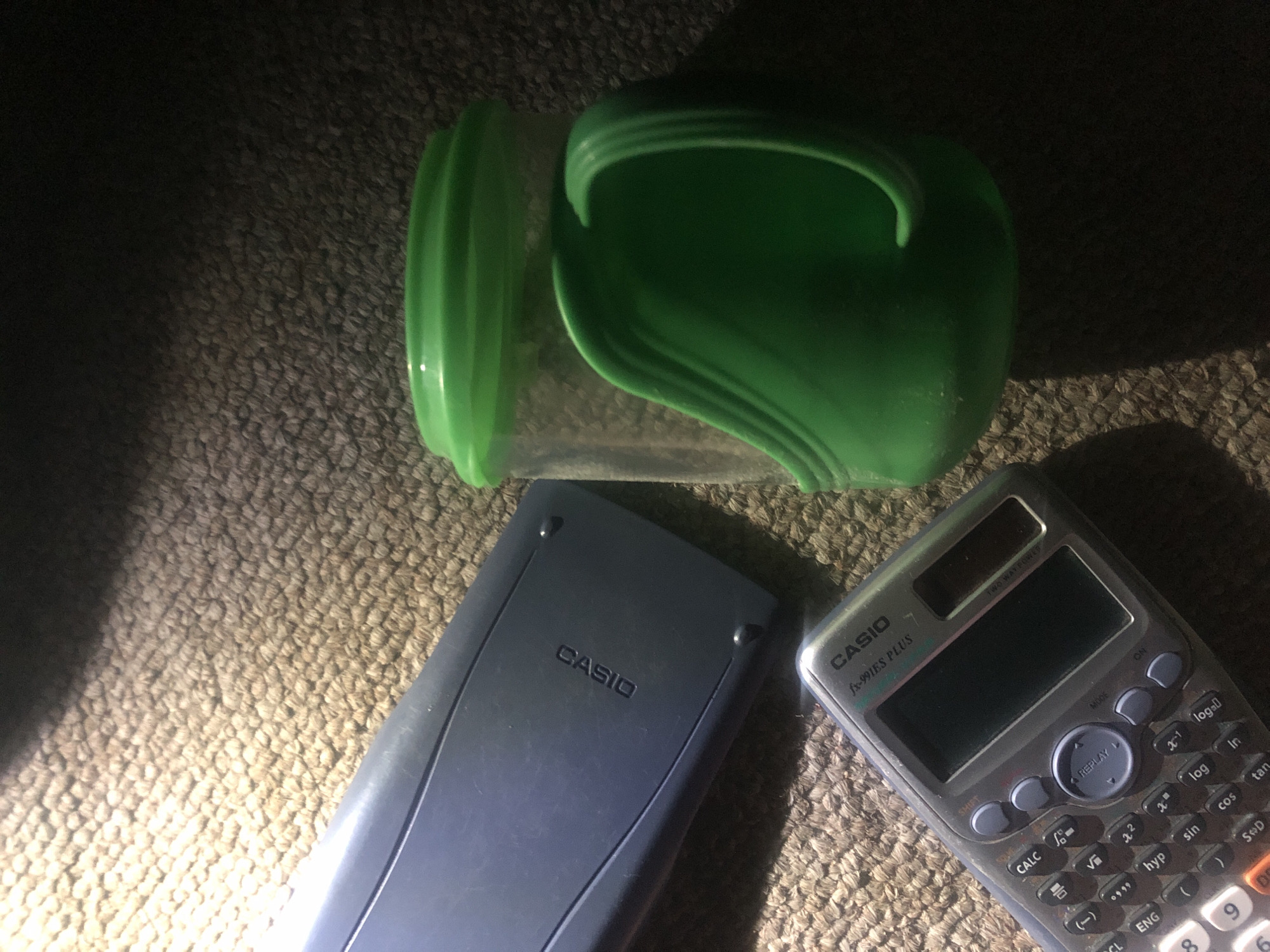 A dark, low-angle, medium shot captures a blue Casio calculator, a darker blue Casio calculator, and a green plastic cup with a handle, all resting on a textured brown carpet. The lighting is dramatic, with a strong light source from the upper left casting shadows and highlighting the items. The blue Casio calculator on the left is lying flat, its surface showing the "CASIO" logo. Below it and slightly to the right is a smaller, silver-grey Casio calculator, angled upwards, displaying a screen and buttons. To the right of the calculators is the green plastic cup, its translucent body visible through the green handle and lid. The cup has a distinct ergonomic handle that curves downwards. The carpet exhibits a coarse weave pattern. The overall impression is one of everyday objects placed together, possibly in a domestic or study setting. The image does not contain information about Jalingo, Nigeria.