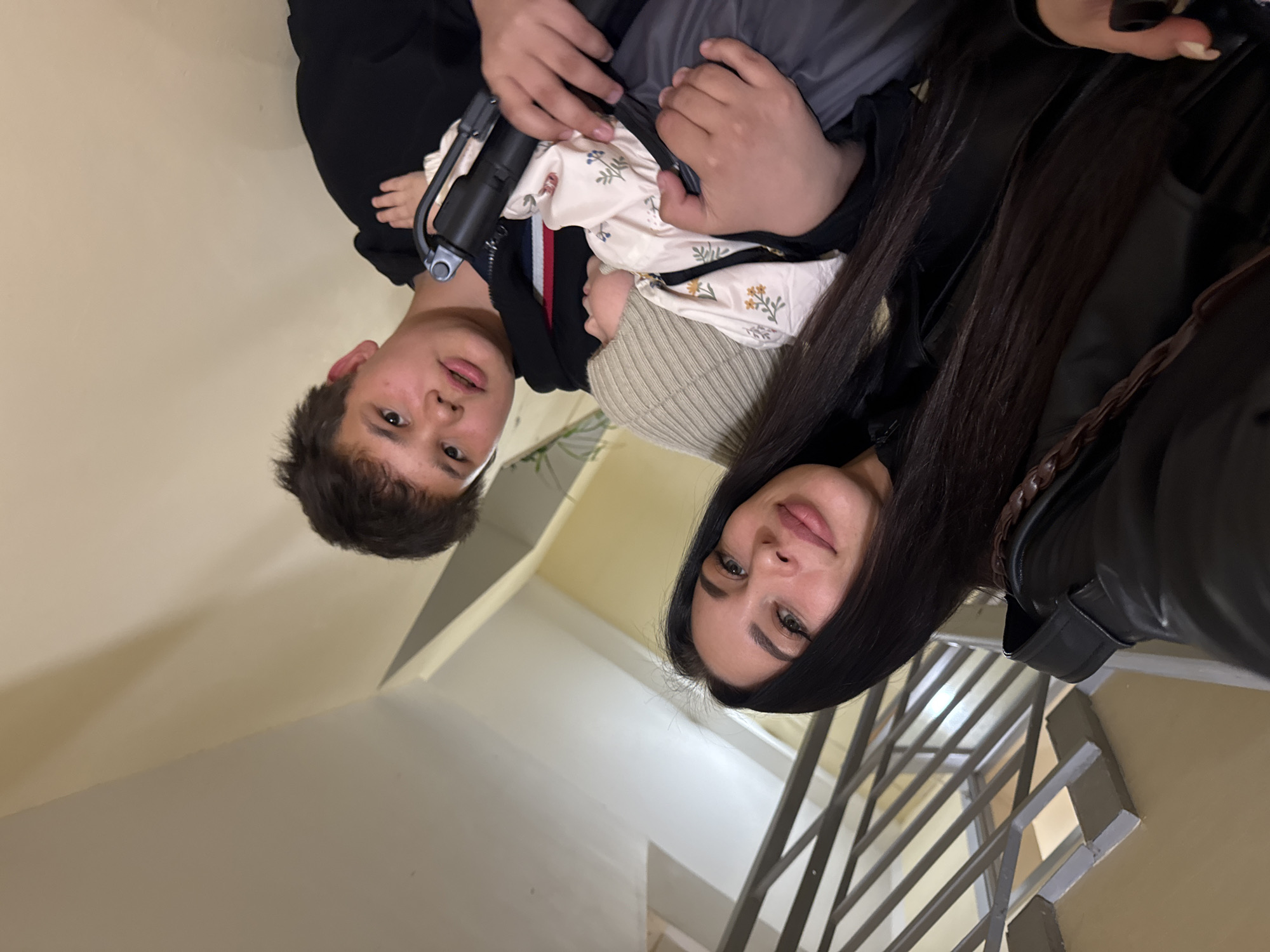 A woman and a boy are in an indoor staircase. The woman has long, dark hair and wears a black leather jacket. The boy is holding a baby, who is wearing a hat and blanket. The staircase has light beige walls and metal railings. Based on the information available, the image was taken in Chaikovsky, Russia.
