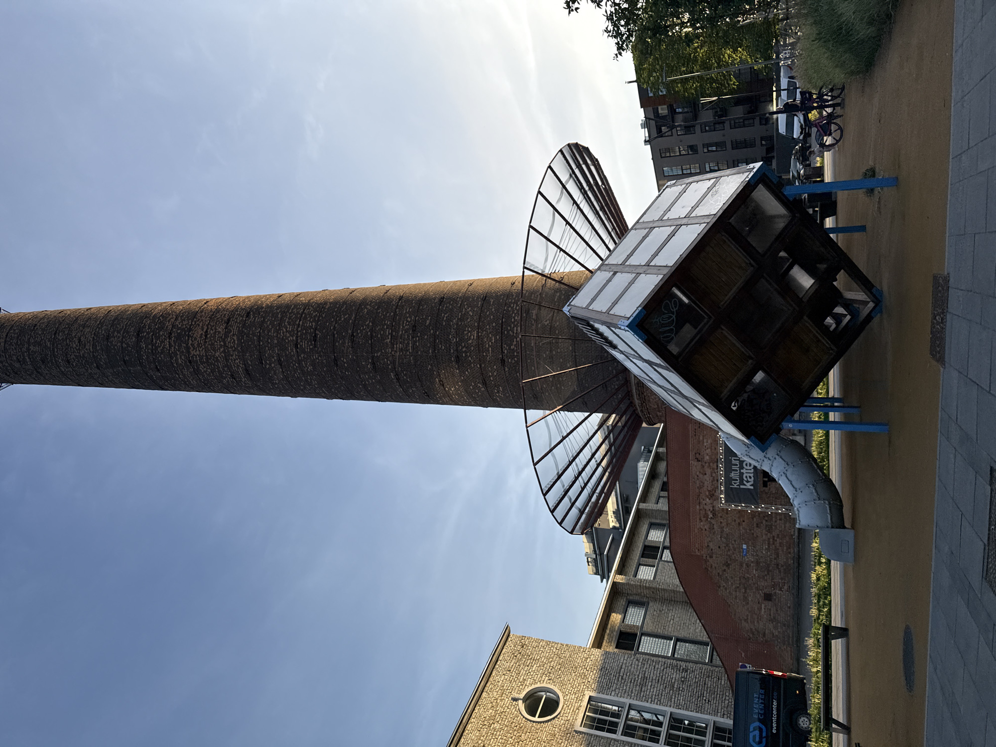 The image depicts an outdoor scene in the city of Tallinn, Estonia, featuring a prominent industrial chimney as the central element. The chimney is tall and constructed from dark bricks, with a visible spiral pattern. Attached to the chimney, at a lower level, is a large, circular structure with a metal frame and glass panels, resembling a satellite dish or a canopy. Below this structure, a cube-shaped installation is suspended, appearing to be made of transparent panels within a wooden frame. This cube is tilted and supported by blue poles. A metal slide extends from the cube downwards towards the ground.
To the left of the chimney and cube installation are several brick buildings. The closest building has a light-colored brick facade with several windows, including a circular window at the top. Adjacent to it is a darker brick building with multiple rectangular windows. In front of these buildings, a black van with "EVENT CENTER" branding is parked. A black bench is positioned on the sandy ground.
In the background, other buildings, including a modern multi-story structure, are visible, along with trees and foliage on the right side of the image. The ground in the foreground is paved with grey tiles, and the area around the play structure is covered in light brown sand. The sky is clear and blue, with some wispy clouds, suggesting it is daytime. Two bicycles are parked in the middle ground. A sign with the text "KULTUURI KATE" is visible on the brick building in the middle ground.