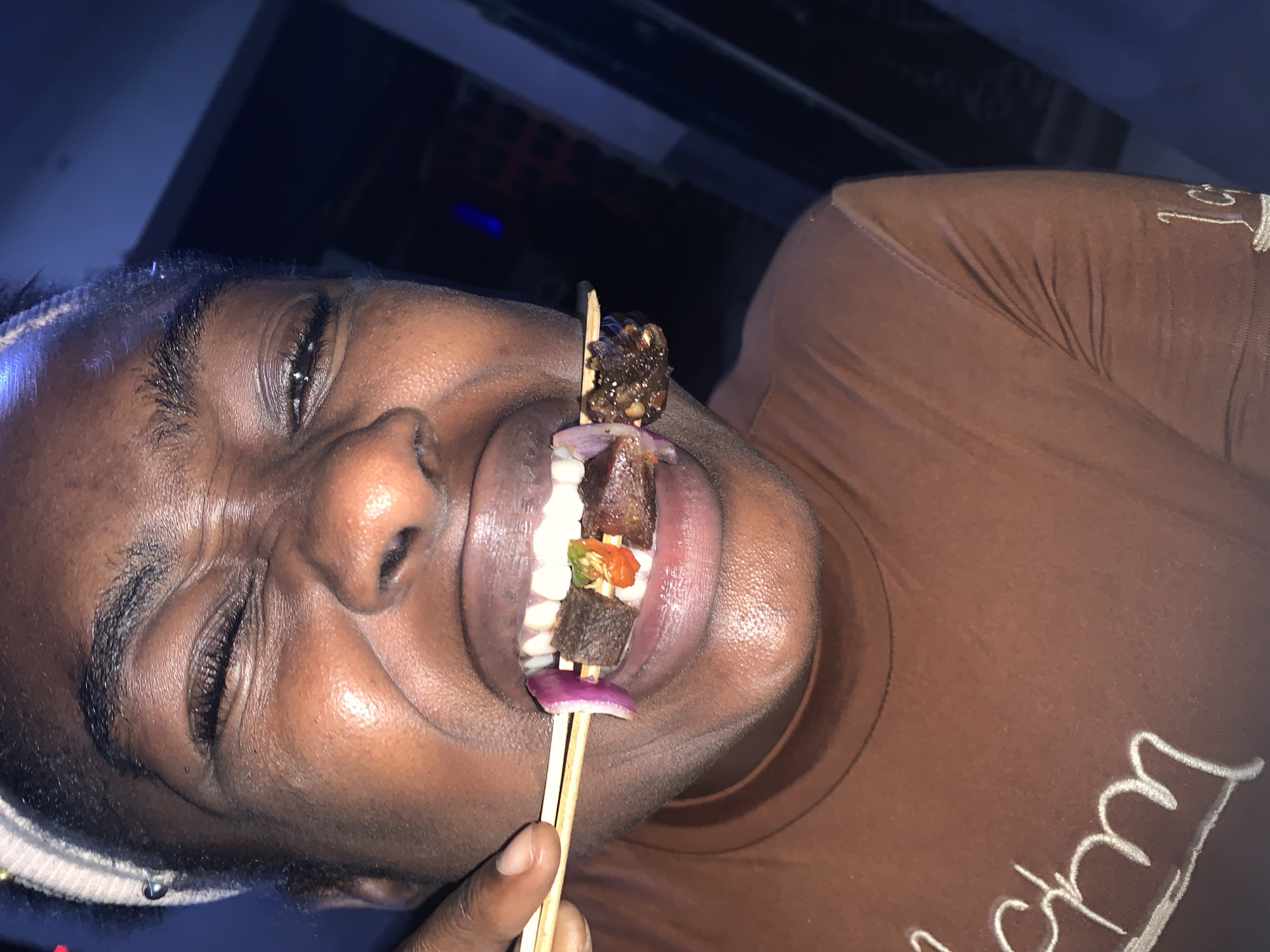 A person with dark skin and a white headband is holding a skewer of grilled meat and vegetables in their mouth. The skewer has a red onion at the bottom, then a piece of meat, a green and red pepper, another piece of meat, and a cluster of smaller grilled pieces on top. The person is wearing a brown t-shirt with white cursive writing on it. The background is dark, with a blue light source visible. The image appears to be taken indoors. Location: likely New Stadium, Nigeria.