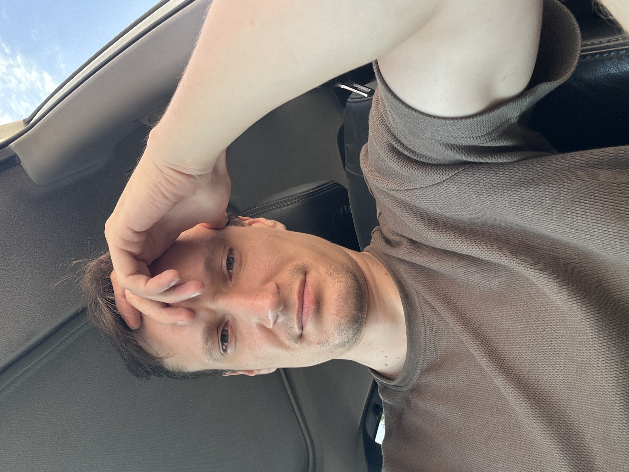 A man, whose right hand is resting on his forehead, is seated in the driver's seat of a vehicle. He is wearing a brown t-shirt. The interior of the car, including the dashboard and a portion of the passenger seat, is visible. The sunroof of the car is open, revealing a bright blue sky with scattered clouds. The scene is outdoors.