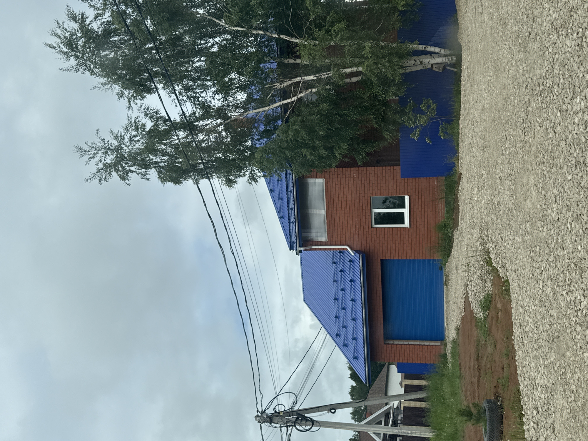 The image displays a red brick house with a distinctive bright blue corrugated metal roof and a matching blue roll-up garage door, situated in a residential area of Perm, Russia, under an overcast sky. The house, possibly two stories, features a white-framed horizontal window on the upper level and a vertical white-framed window on the lower right facade. A white downspout is visible on the right side of the building. In front of the garage, a light-colored gravel driveway curves across the foreground, interspersed with patches of bare earth and sparse green vegetation. To the left of the driveway, a small evergreen plant grows within an old tire. A concrete utility pole with numerous black power lines stands prominently on the far left, with lines extending across the upper portion of the frame. Several tall, leafy trees, including multiple birch trees with white bark, partially obscure the right side of the house and its adjacent blue corrugated metal fence. A birdhouse is attached to one of the birch trees. In the distant background on the left, parts of another house with a light-colored gable roof are visible. No people or animals are present in the scene.