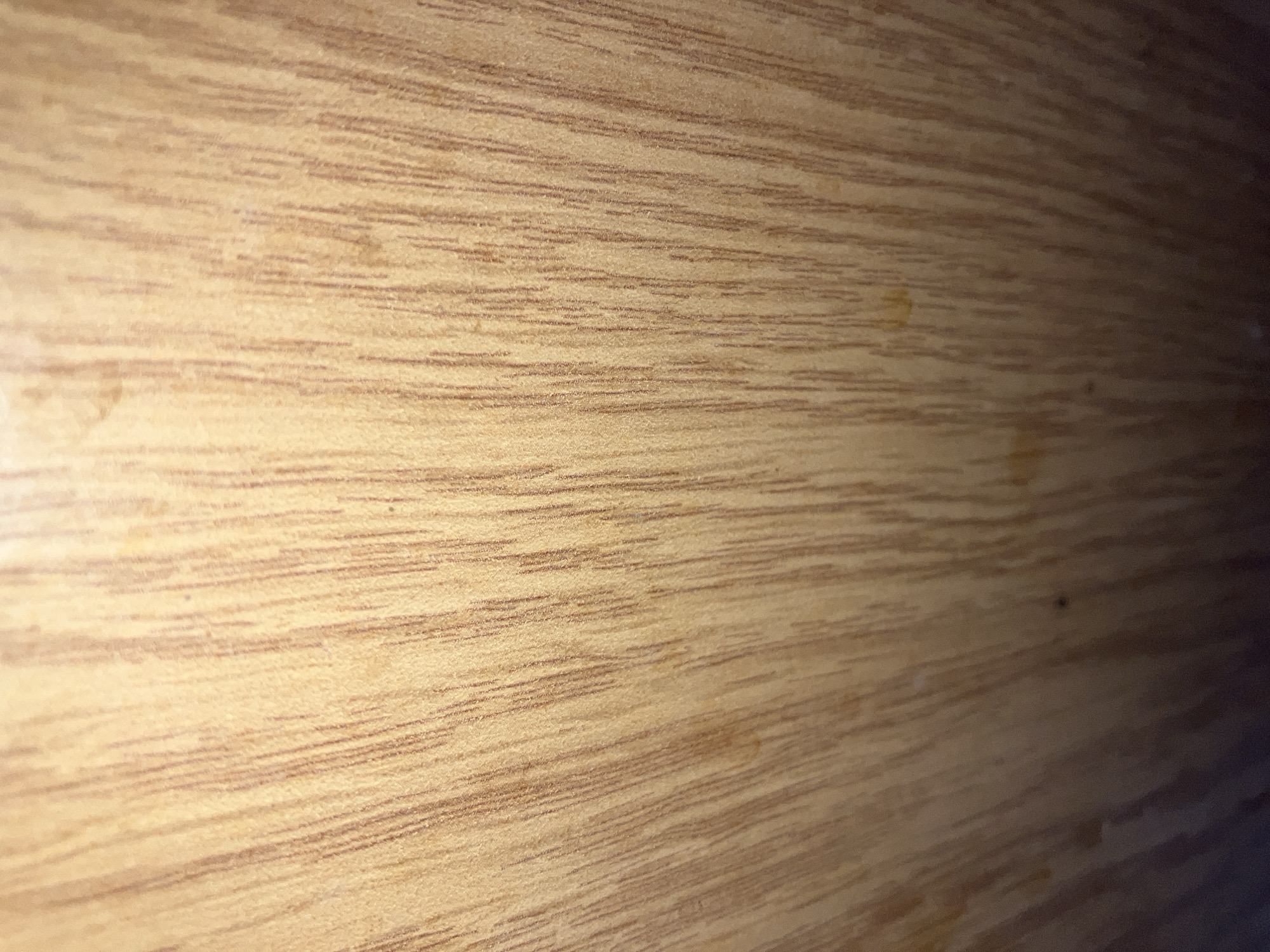 The image is a close-up, textured shot of a light-colored wooden surface. The wood grain is prominently displayed with vertical, thin, dark brown lines running parallel to each other. There are some lighter, irregular splotches and darker spots scattered across the surface, suggesting natural variations in the wood or minor imperfections. The lighting appears to be coming from above and slightly to the left, casting a subtle shadow that creates a gradient from a lighter top to a darker bottom. The image lacks any specific identifiable landmarks or context that would allow for localization in Kyiv, Ukraine. There are no people, objects, actions, or interactions depicted.