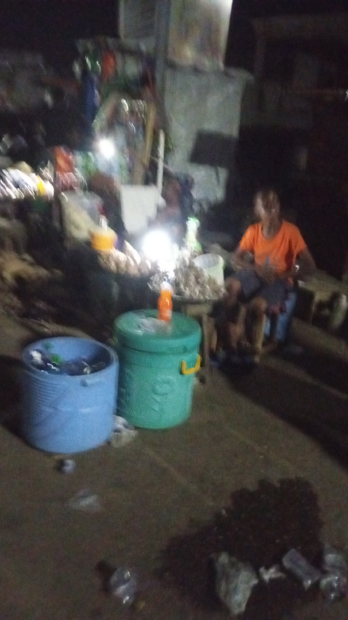 The image presents a blurry, dimly lit outdoor scene at night, likely depicting a market stall or roadside vendor in Lokoja, Nigeria.
In the foreground, two large plastic containers are prominent: a light blue open bucket on the left, containing some miscellaneous items, and a closed green cooler with a yellow handle, on top of which rests a bright orange bottle. The ground appears uneven and littered with debris, including what looks like discarded plastic bottles and other refuse.
Towards the right, a person wearing an orange shirt is seated, facing left, seemingly attending to their wares. They are positioned behind a surface, possibly a table or stall, laden with numerous light-colored items, which are difficult to identify precisely due to the blurriness but could be dried produce or other goods. A very bright, circular artificial light source intensely illuminates these items and the immediate vicinity, suggesting its necessity for operations in the dark. The background is indistinct, showing blurry structures that imply a built environment.
The scene captures a functional commercial activity taking place after dark, heavily reliant on artificial lighting. No visible text is discernible.