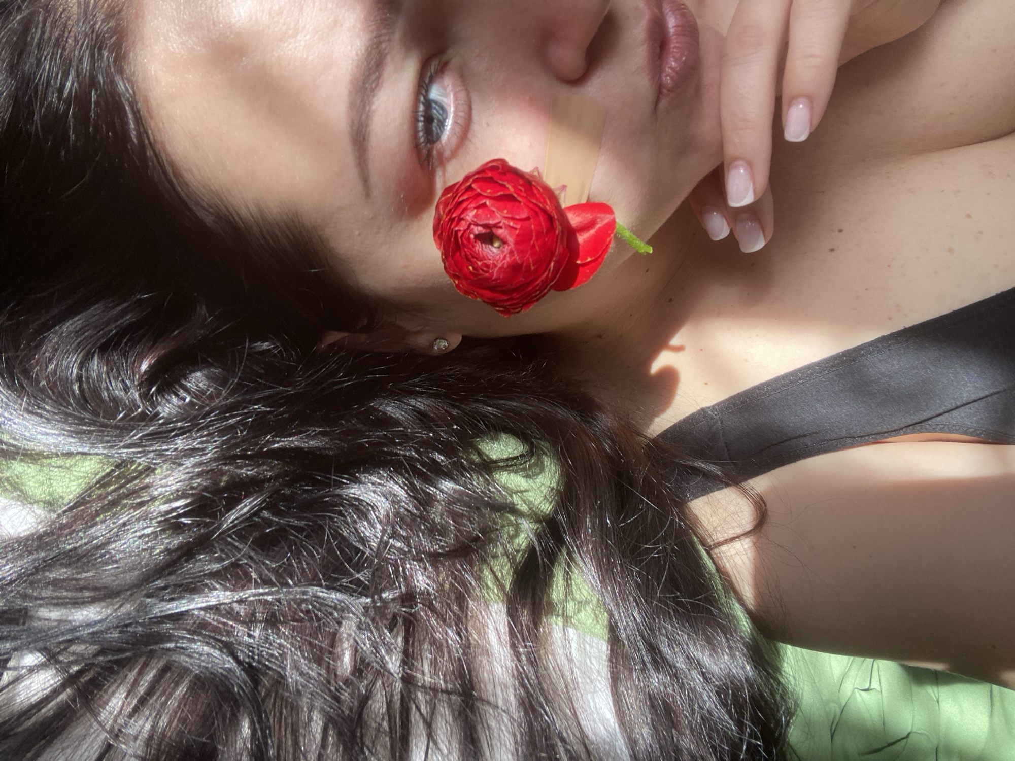 A close-up, high-angle shot captures a person, likely female, lying down with their head tilted upwards, possibly in Sochi, Russia. Their face is partially visible, featuring a light-toned complexion, a clear blue eye looking upwards, and a small diamond stud earring in their visible ear. Long, dark, wavy hair cascades around their head, showing highlights from the strong, natural sunlight.
A vibrant red ranunculus flower with multiple delicate petals is centrally placed on their cheek, near the nose, almost appearing to emerge from the skin. A small, beige rectangular bandage is visible on the cheek, positioned just above and to the right of the flower.
One hand, with neatly manicured nails featuring white tips (French manicure), rests near their neck and lower face. The person wears a black top or bra, with a strap visible. Strong, directional sunlight illuminates the scene, casting distinct shadows across their face, body, and hair, and highlighting the textures. A soft, light green fabric is visible in the background, contrasting with the dark hair and black attire. The overall mood is serene and natural, captured in bright daytime conditions. No text is visible in the image.