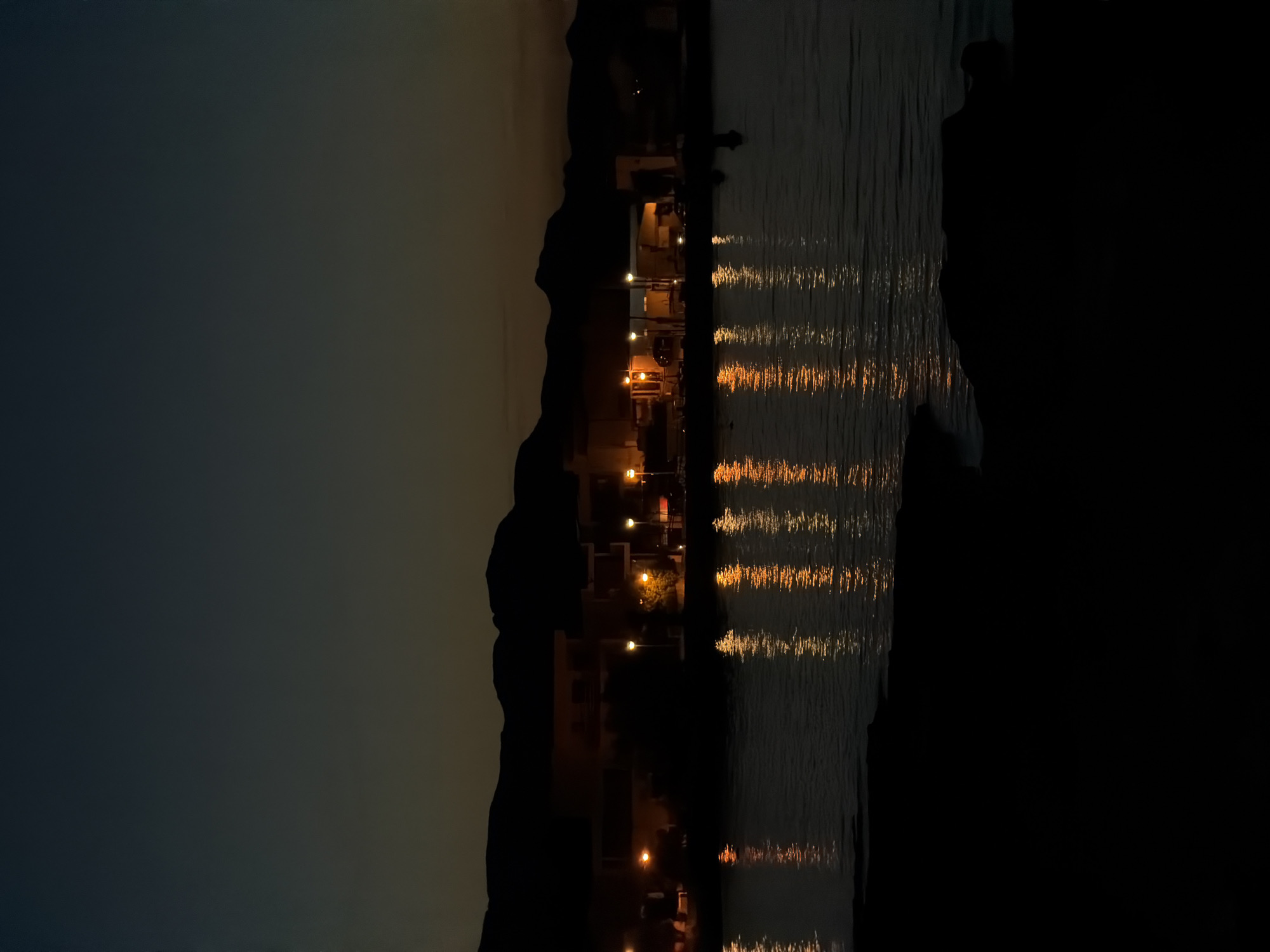 The image shows a nighttime scene in Alcúdia, Spain. The sky transitions from dark to light, suggesting either dusk or dawn. Along the shoreline, a silhouette of hills or mountains is visible. Lights from buildings, likely homes or businesses, are present and their reflections create shimmering, horizontal patterns on the water's surface. A dark mass may represent a standing person. A large area of blackness dominates the right portion of the picture.