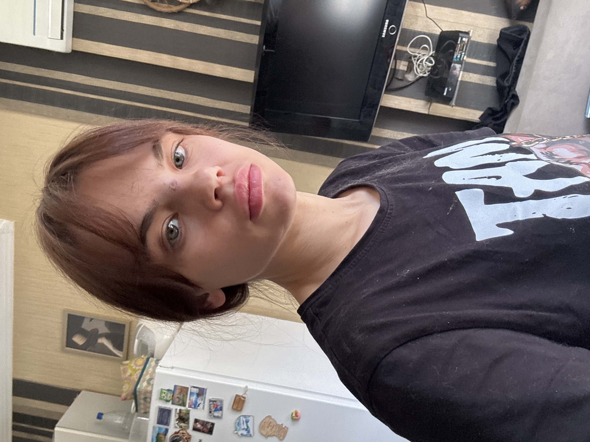 A young person with fair skin, shoulder-length brown hair with bangs, and light-colored eyes is visible in the image. They are wearing a black t-shirt with a large white design. The person is facing the camera with a slight downward tilt.
Behind the person is a cluttered environment. A flat-screen television is mounted on a wall with horizontal stripes. A small electronic device and tangled wires are visible near the television. A white refrigerator is also present, adorned with magnets and pictures. A framed picture hangs on the wall near the refrigerator.
The location is not discernible as being Rostov-on-Don, Russia from the visible details.