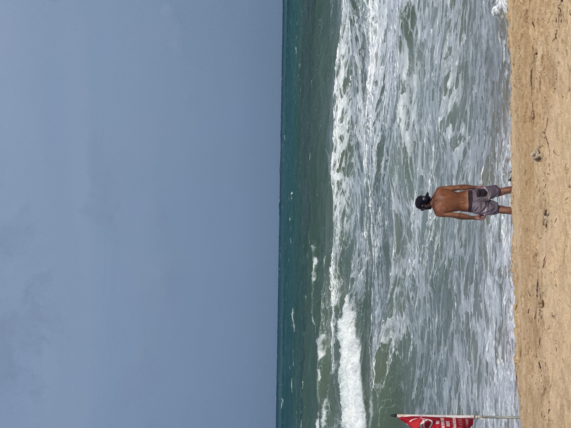 A man stands on a sandy beach, facing a turbulent sea under a gray, overcast sky. He is shirtless, wearing gray swim trunks and a black baseball cap. A red flag with white lettering, a warning sign, is planted to his left, its text partially obscured. The waves are crashing onto the shore, creating white foam. The ocean stretches to the horizon, meeting the sky in a seamless blend of blue and gray. The scene appears to be located on a beach in Rawai, Thailand, known for its beautiful but sometimes rough seas.