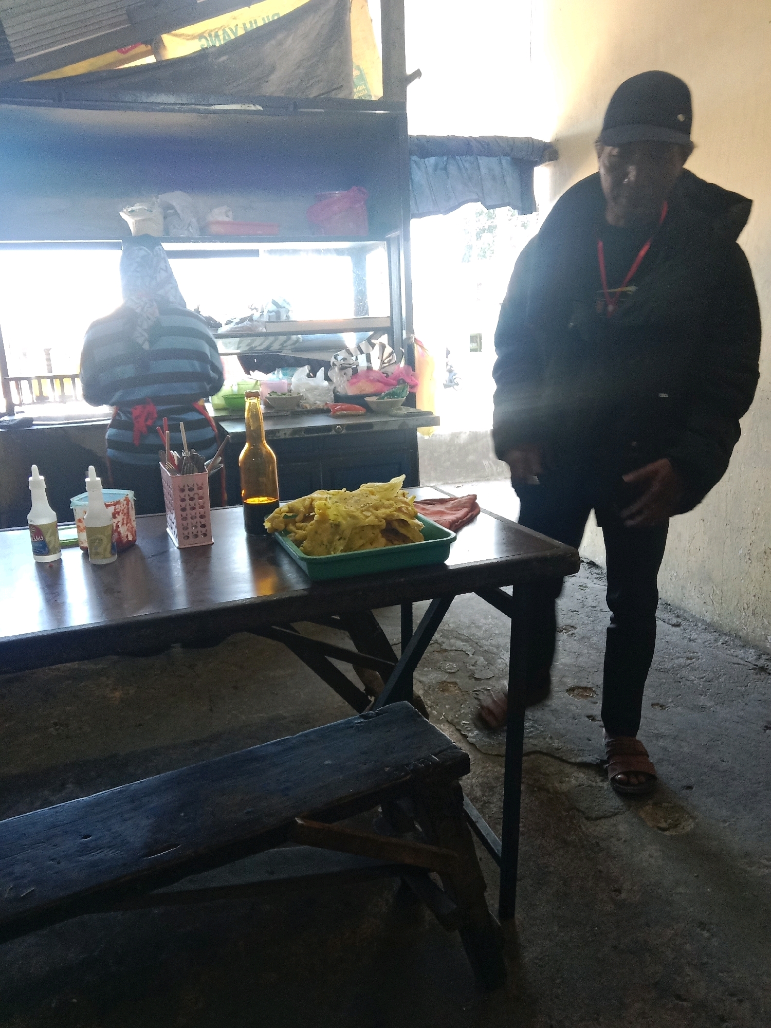 A man in a dark jacket and cap stands near a table laden with food and condiments in an outdoor food stall in Candiyasan, Indonesia. To his left, a person wearing a striped shirt and headscarf is seen from behind, attending to the stall. The table holds two white squeeze bottles, a pink cutlery holder with chopsticks, a brown glass bottle, and a green container filled with fried food. The stall appears to be open-air with a concrete floor and a corrugated metal roof overhead. The background features a counter with various items and a bright, hazy view of the outdoors.