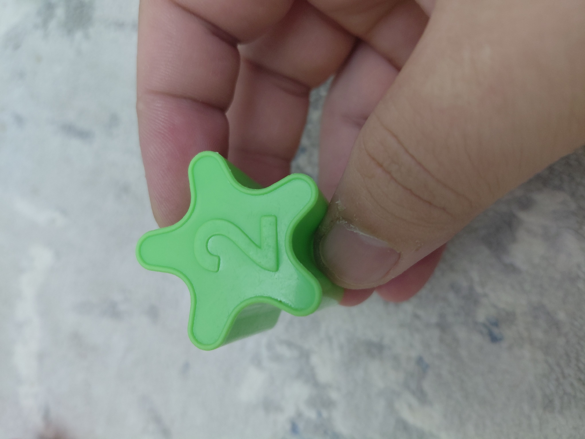 A bright green, star-shaped plastic object with the number "2" embossed on its surface is held between the thumb and forefinger of a person's hand. The background consists of a light-colored, textured surface, possibly a carpet or fabric. The image does not provide any information about the location in Aktau, Kazakhstan.