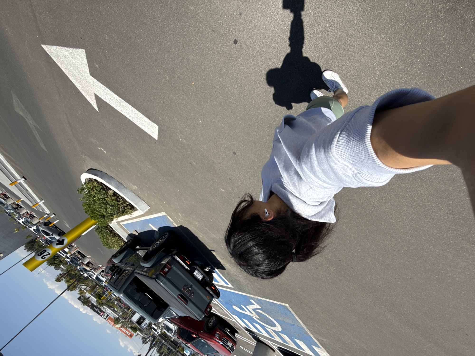 The image depicts an outdoor commercial parking lot under clear, sunny conditions. The scene is captured from a tilted, upward-angled perspective, indicative of a selfie.
In the foreground, the upper torso and head of an individual are visible, facing away from the camera and slightly to the right. This person has dark hair tied in a low ponytail, wears a light grey hooded sweatshirt, light green shorts, and white sneakers. A white earbud is inserted into their right ear. Their right forearm and hand, extending the camera, are visible in the lower right portion of the frame. A distinct shadow of a person, likely the photographer, is cast onto the asphalt surface to the right.
The mid-ground features a dark grey Jeep Gladiator pickup truck parked in an accessible space, indicated by a blue wheelchair symbol and the word "DISCAPACITADOS" painted on the asphalt. The truck's license plate, partially visible, reads "2-5500A". A red SUV is parked immediately to the left of the Jeep. Adjacent to these vehicles, a tall, bright yellow pole with a circular sign displaying the number "26" and a black left-pointing arrow stands next to a small, circular concrete planter containing green shrubs. A large white arrow is painted on the asphalt, pointing diagonally upward and to the right.
The background shows multiple rows of parked vehicles and the exterior of a commercial shopping center. Retail signage for "LA PLACENTERA," "99," and "EXTRA" are discernible. Overhead utility cables or power lines traverse the blue sky, which has minimal white cloud cover. Palm trees and other foliage line the distant horizon.
This scene is located in a commercial area within the city of Puebla, Mexico, evidenced by the specific retail chains and Spanish-language parking markings.