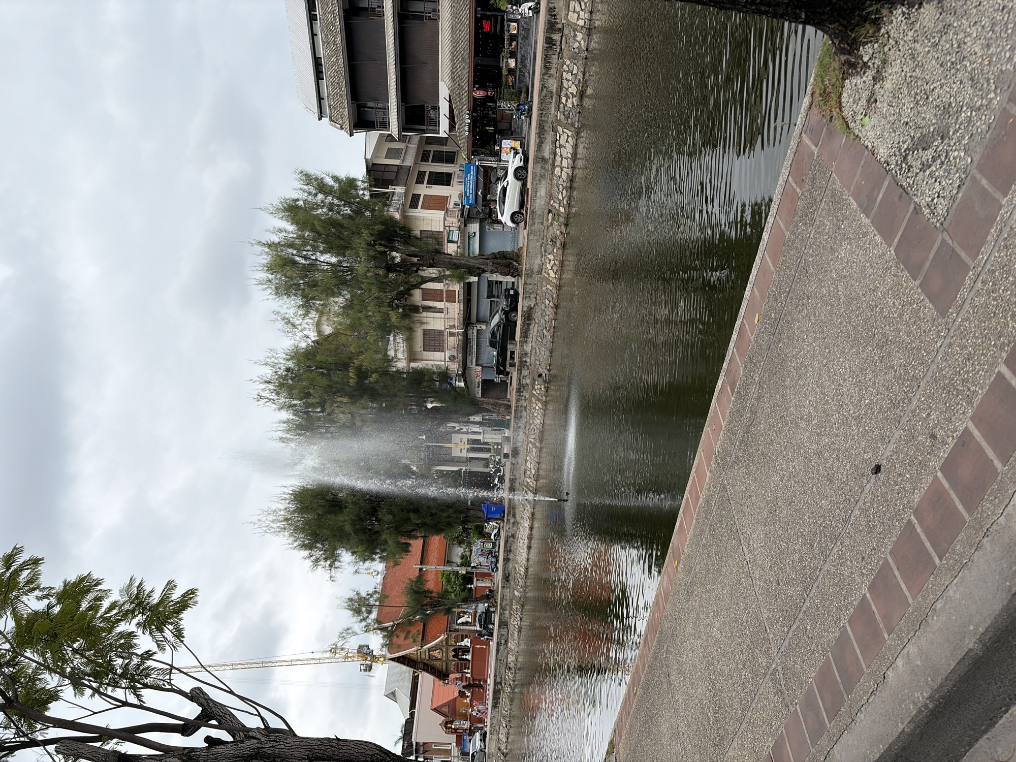 This is a daytime, eye-level, medium shot of a canal in Chiang Mai City Municipality, Thailand. A fountain is visible in the canal, spraying water upwards. The canal is lined with a stone wall and a paved walkway. Beyond the canal, there are buildings, trees, and parked vehicles. One building appears to be a temple with a red roof, while others are more modern commercial structures. A yellow construction crane is visible in the distance. The sky is overcast. There are no people clearly visible in the image.
