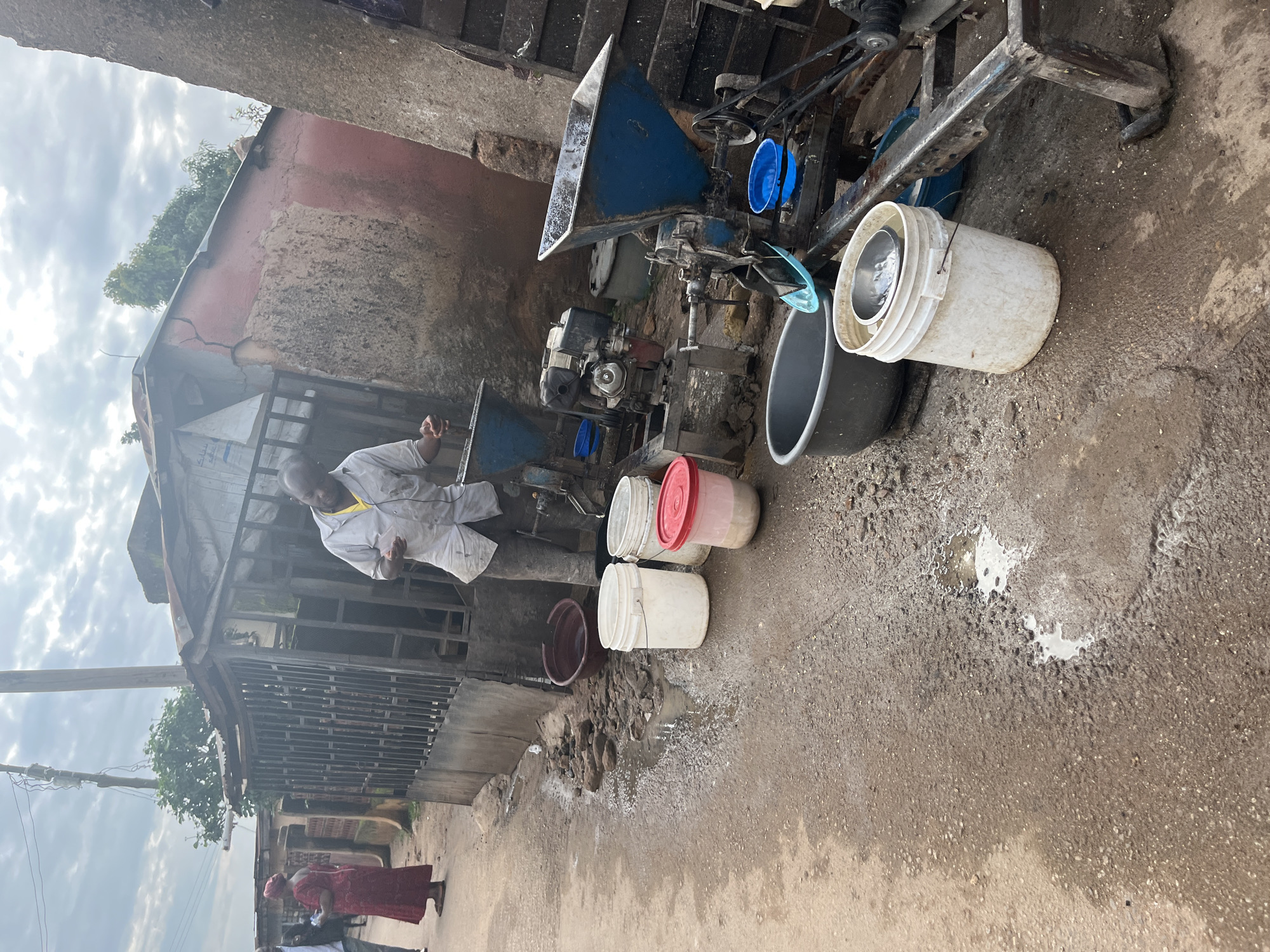 The image depicts an outdoor scene in the city of Jos, Nigeria, characterized by a dusty, unpaved street with puddles of water. In the foreground, several white plastic buckets, a red-lidded container, and a dark brown basin are arranged near some industrial equipment. This equipment appears to be a milling or grinding machine, powered by a small engine, with a blue hopper and various belts and pulleys. A man, wearing a light-colored shirt and pants, stands by the machinery, seemingly engaged in its operation. To the left of the scene, two women, one in a red dress and the other in a darker garment, are walking away from the camera along the street. A metal gate and a dilapidated building with a corrugated metal roof form the backdrop. The overall environment suggests a commercial or industrial area operating in a somewhat rustic setting.