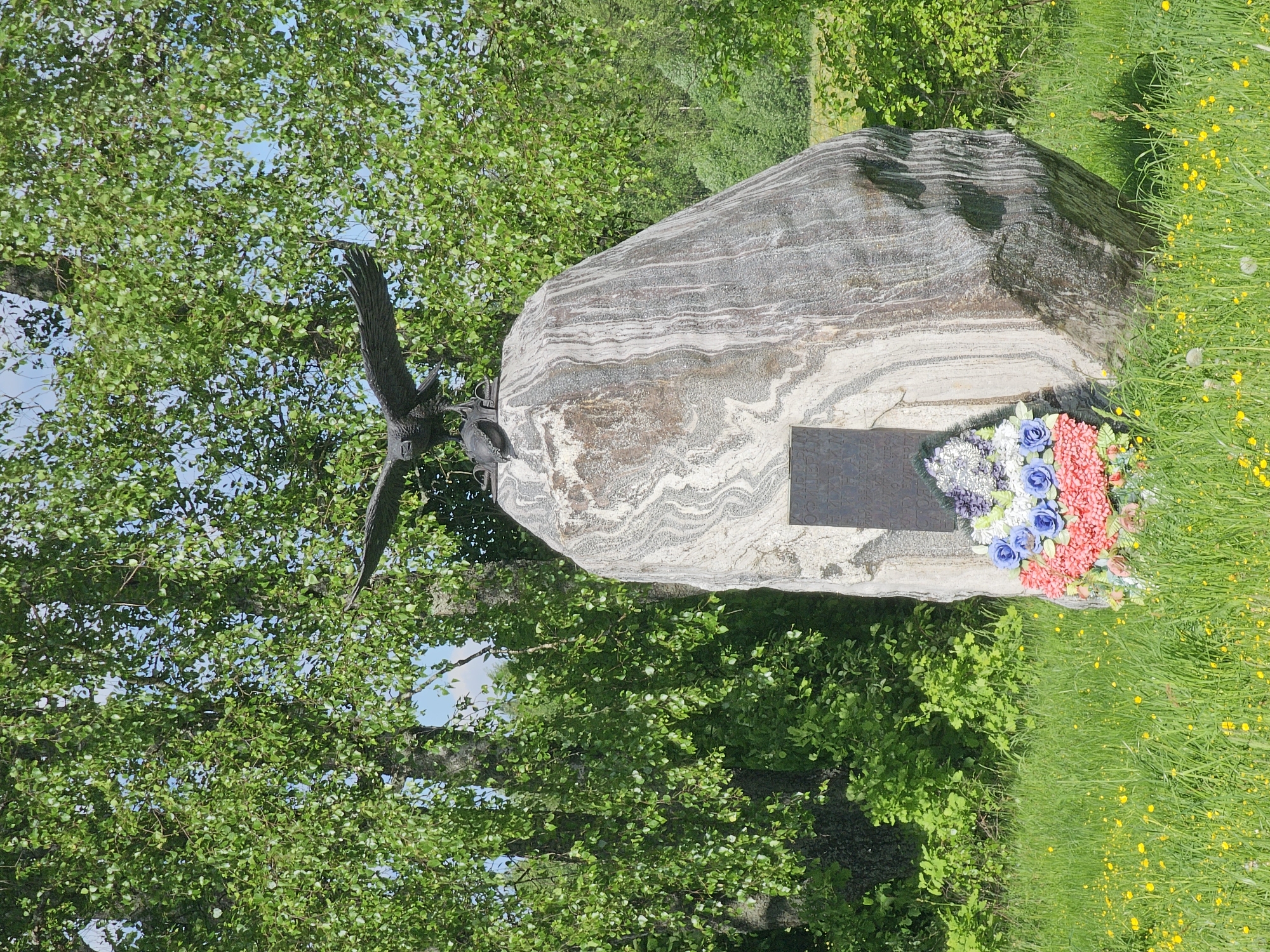 The image depicts a memorial in Borodino, Russia, possibly dedicated to military or historical events. A large, natural stone serves as the central element, featuring layered geological patterns. A metal sculpture of an eagle is mounted on the stone. A memorial plaque is attached to the stone, and a colorful wreath of artificial flowers rests nearby. The monument is situated in a grassy area surrounded by trees.