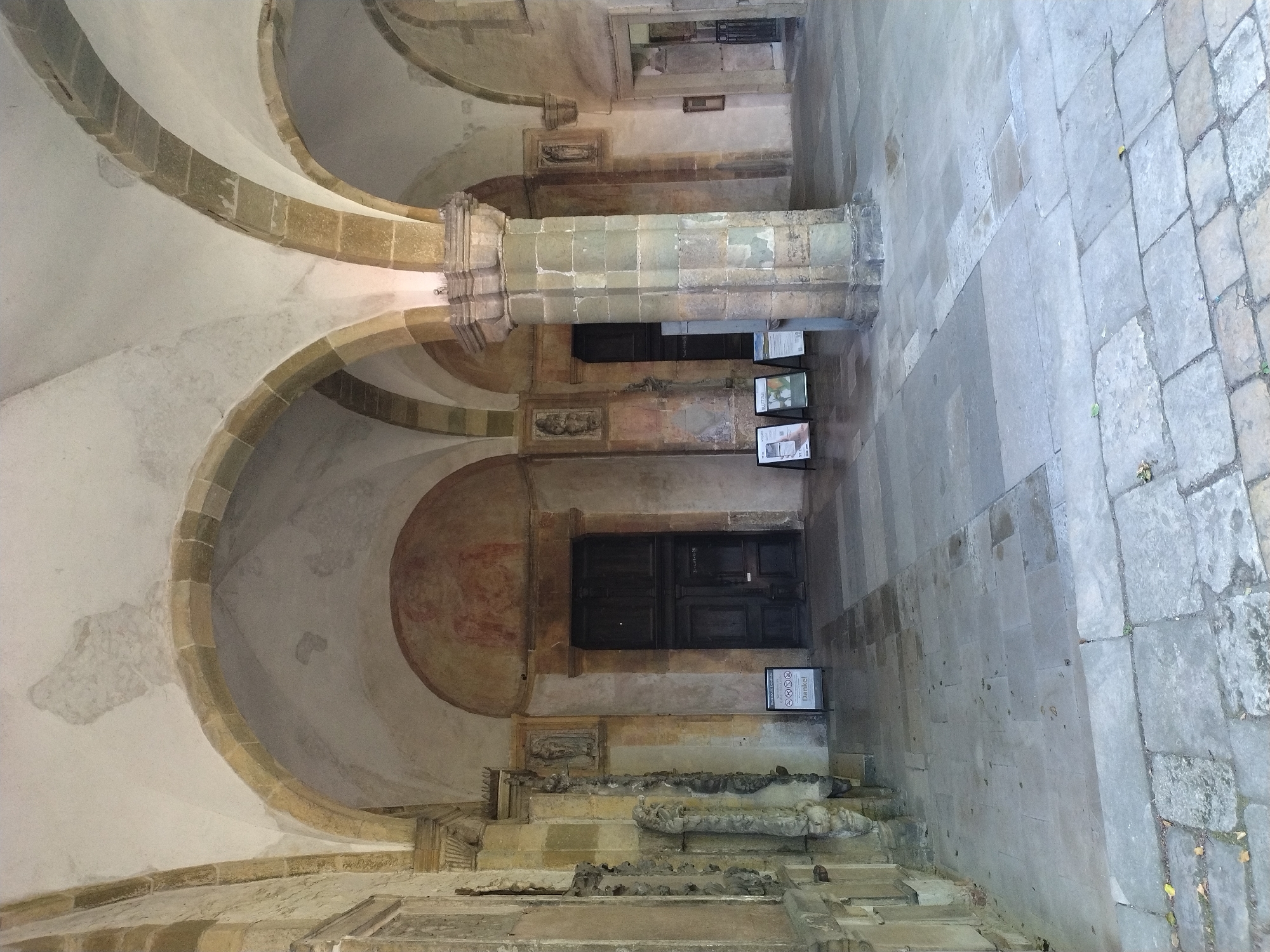 The image shows a view of the St. Emmeram's Abbey in Regensburg, Germany. The scene is an outdoor cloister area with stone-paved flooring and arched walkways. In the background, there is a large wooden door set within an arched entryway, with faint paintings above it. Several informational stands with signs are visible in front of the entryway. The architecture features stone columns, arches, and decorative elements on the walls. The lighting suggests it is daytime, with shadows indicating the presence of overhead structures. The scene is devoid of any visible people or animals.