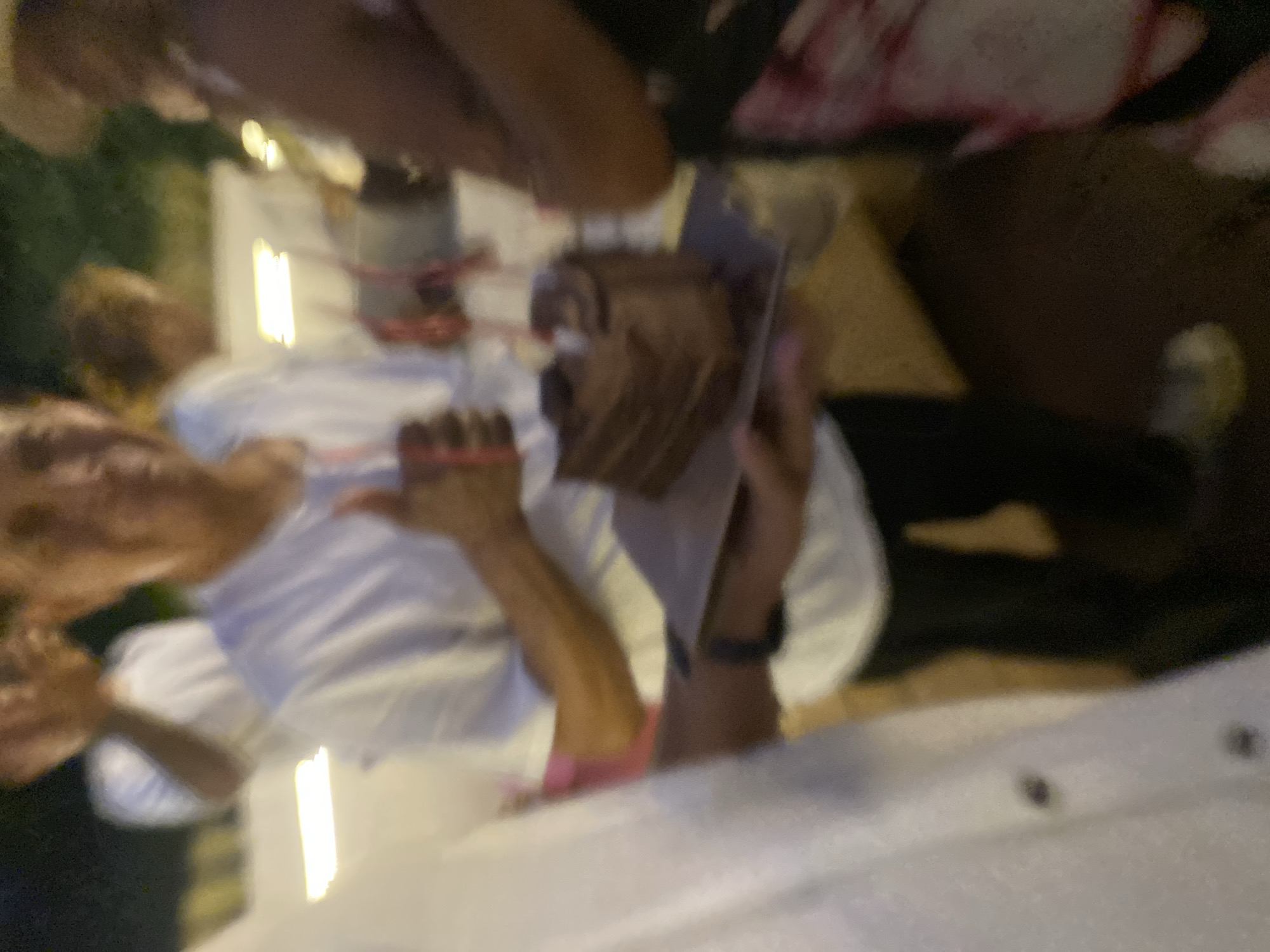 This media file captures a moment at a gathering, likely a party or celebration, with several people present. In the foreground, a person is holding a small, chocolate-colored cake, possibly a cupcake, decorated with what appears to be frosting and perhaps sprinkles. Another person, wearing a white shirt, is visible in the background, along with parts of other individuals. The scene suggests an indoor or semi-outdoor setting with ambient lighting. The overall image is somewhat blurry and tilted, making it difficult to discern fine details of the individuals or their attire. The context of Nusa Penida, Indonesia, implies a tropical or coastal location, though specific environmental cues are not prominent in the image. The event appears to be casual and celebratory, given the presence of the cake.