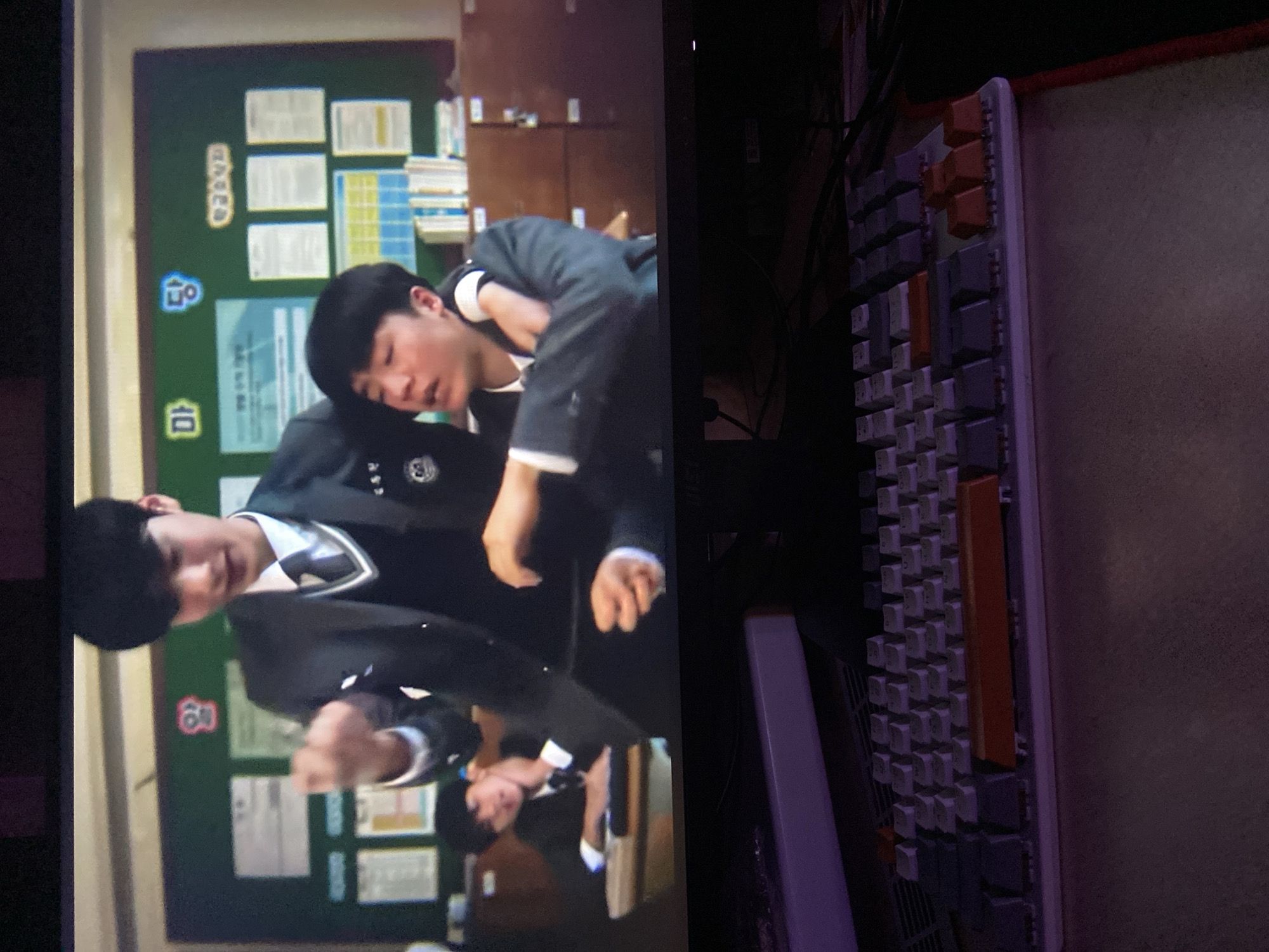 In the foreground, a portion of a white and purple mechanical keyboard is visible, with several keys illuminated with an orange tint. The background shows a television screen displaying a scene from a Korean drama. Two young males in school uniforms are depicted in a classroom setting. One is standing and gesturing with his right hand, while the other is seated, with his arm around the standing male's shoulder. The classroom has a green chalkboard with posters and papers displayed on it, and wooden cabinets filled with books. The scene is consistent with a school environment in South Korea, not Beloretsk, Russia.