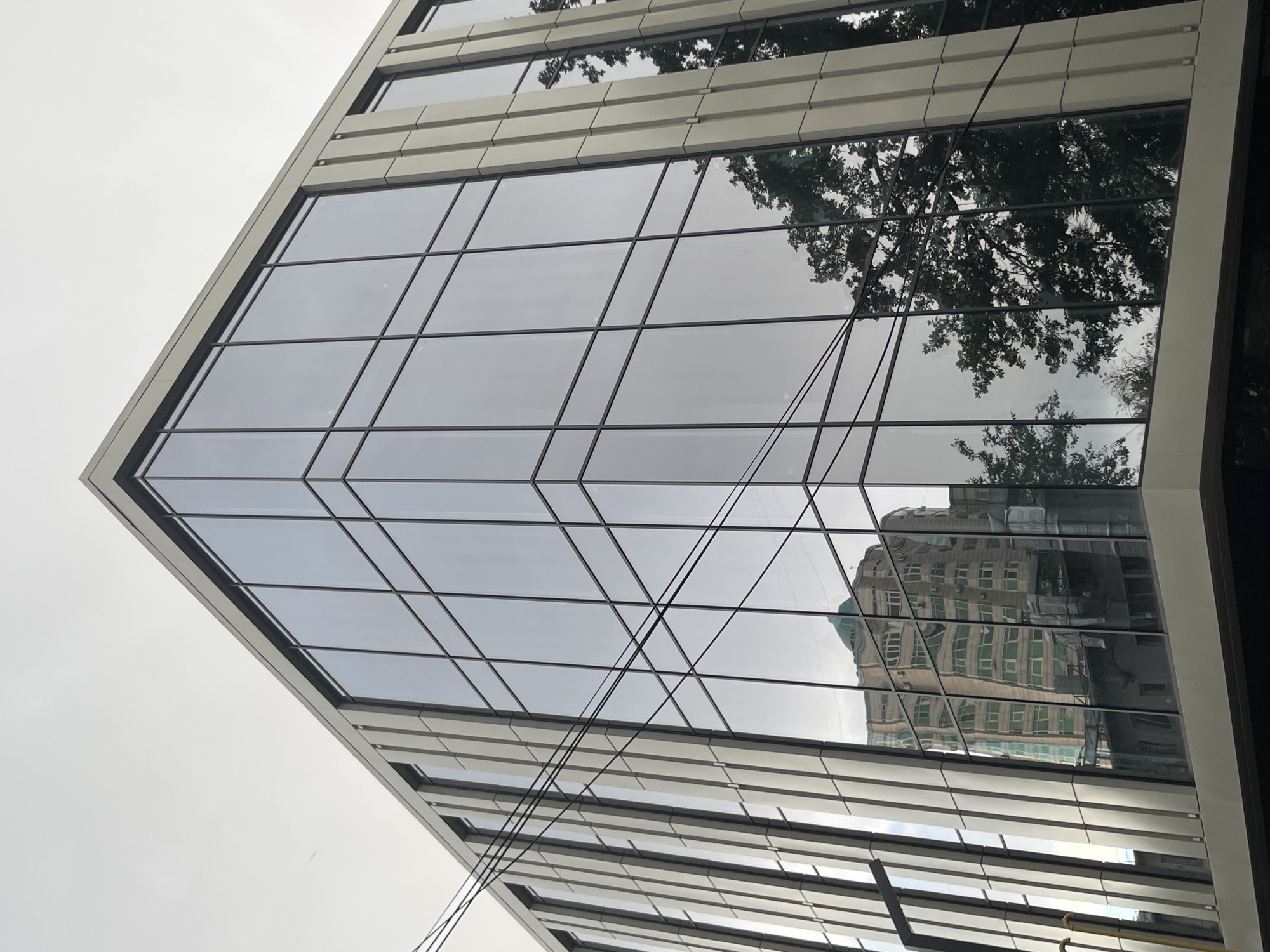 This media file shows a modern building with a glass facade, viewed from a low angle looking upwards. The building has a contemporary architectural style with clean lines and a reflective surface. The glass panels are arranged in a grid pattern, with thin dark frames separating them. Some panels reflect the sky, which appears overcast, suggesting it is either morning or late afternoon with diffused light.
The reflection in the glass reveals a large tree with dense foliage, its branches stretching across the upper portion of the facade. Below the tree's reflection, a building with multiple floors and visible windows is reflected. This reflected building appears to be older, with a more traditional architectural style, featuring a light-colored facade and dark window frames. The contrast between the modern glass building and the reflection of the older building creates an interesting visual juxtaposition.
Thin black wires or cables are visible crisscrossing in front of the glass facade, adding a sense of depth and perhaps indicating some functional element of the building or its surroundings. The overall impression is one of urban architecture, where new construction interacts with its environment and existing structures through reflection. The scene suggests a location in a city, possibly Almaty, Kazakhstan, as indicated by the provided context, where modern office buildings or residential complexes are common. There is no visible text in the image. The weather appears to be cloudy or overcast, and the time of day is indeterminate due to the lighting conditions.