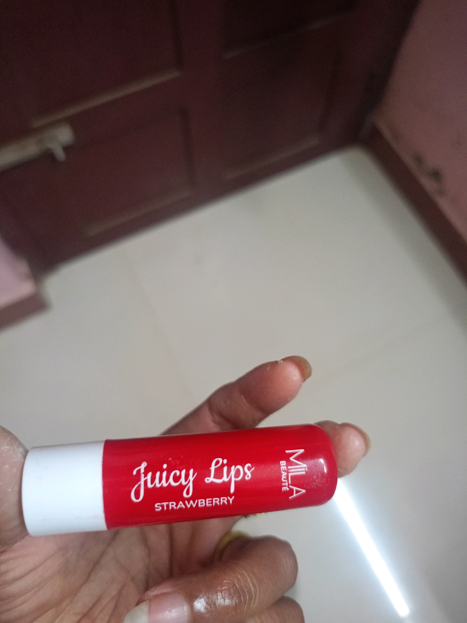 A close-up, eye-level, interior shot shows a hand holding a red and white tube of "Juicy Lips Strawberry" lip balm, with the brand name "Mila Beaute" visible. The background features a blurred, dark red wooden door and a light-colored floor. The image appears to be taken in a residential setting in Chenkavila, India.