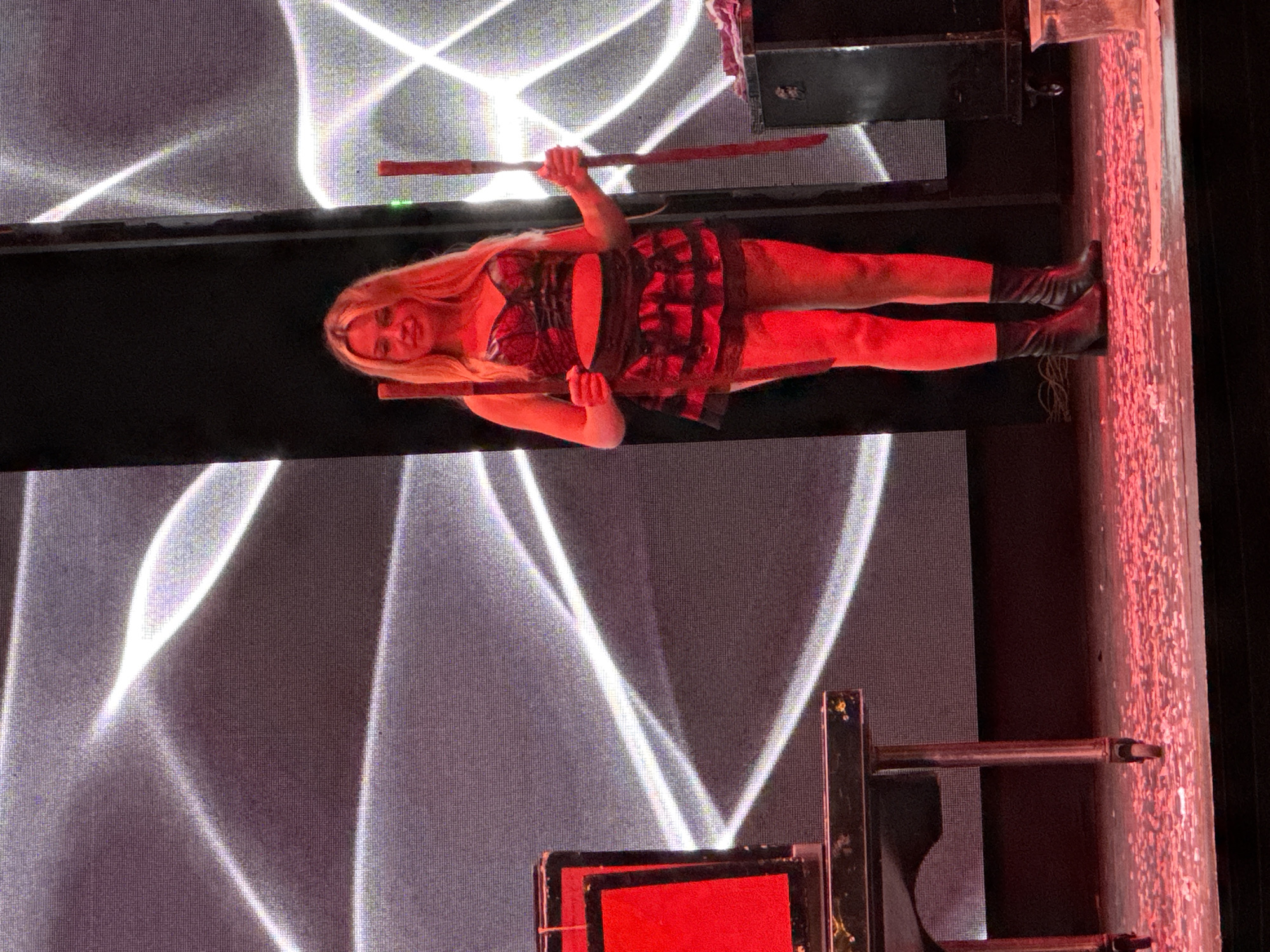 A woman stands on a stage in Manavgat, Turkey, holding two wooden rods. She is wearing a black, sequined top and a matching, ruffled skirt, along with black ankle boots. Her blonde hair is down, and she is illuminated by red stage lighting. In front of her is a black table with a red object on it, and to her right, a black cabinet. The stage floor is covered in red confetti. The background features a large screen displaying a pattern of white lines.