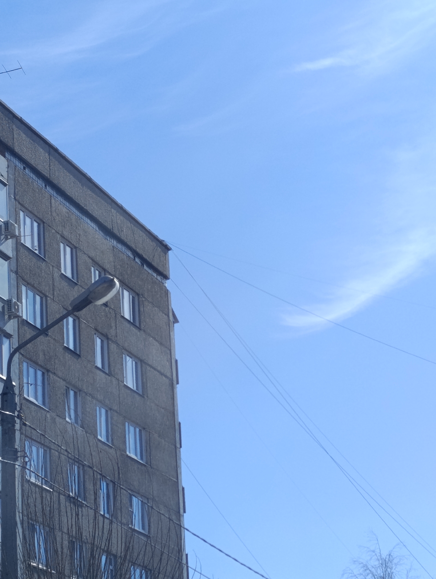The image shows the corner of a grey, multi-story building under a blue sky, likely taken in Krasnoyarsk, Russia, judging by the typical Soviet-era architecture. The building has rows of windows, some with air conditioning units. A street lamp stands in front, obscured by bare tree branches. Power lines extend across the sky. No people are visible.