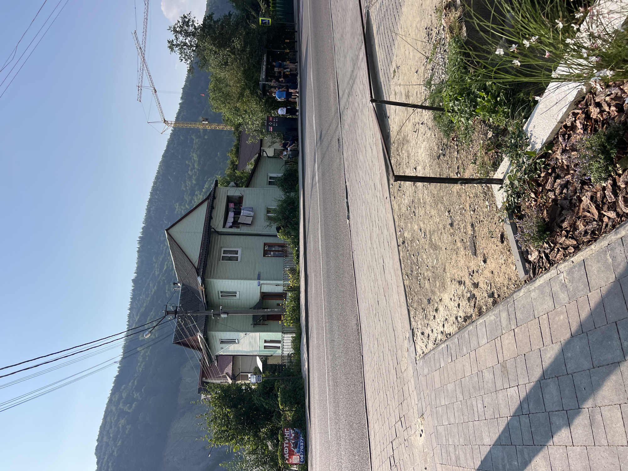 The image depicts a street scene in Yaremche, Ukraine, on a clear, sunny day. In the foreground, a paved sidewalk transitions to a dirt area with landscaping, featuring mulch and green plants. A road runs parallel to the sidewalk, with white lane markings.
Across the road, several buildings are visible. A light green, two-story house with white trim and dark green shutters is prominent. Clothes are hanging on a clothesline outside one of the upper windows. To the left of this house, a red billboard with the word "РАФТИНГ" (rafting) and a phone number is displayed. Further down the street, a bus stop with a sign reading "АВТОСТАНЦІЯ вул. Свободи" (Bus station, Svobody Street) is situated.
Several people are gathered at the bus stop, some sitting and others standing. Two yellow construction cranes are visible in the background, positioned near a wooded hillside. The environment is characterized by a mountainous landscape covered in dense green trees under a bright blue sky. Power lines stretch across the upper portion of the image.