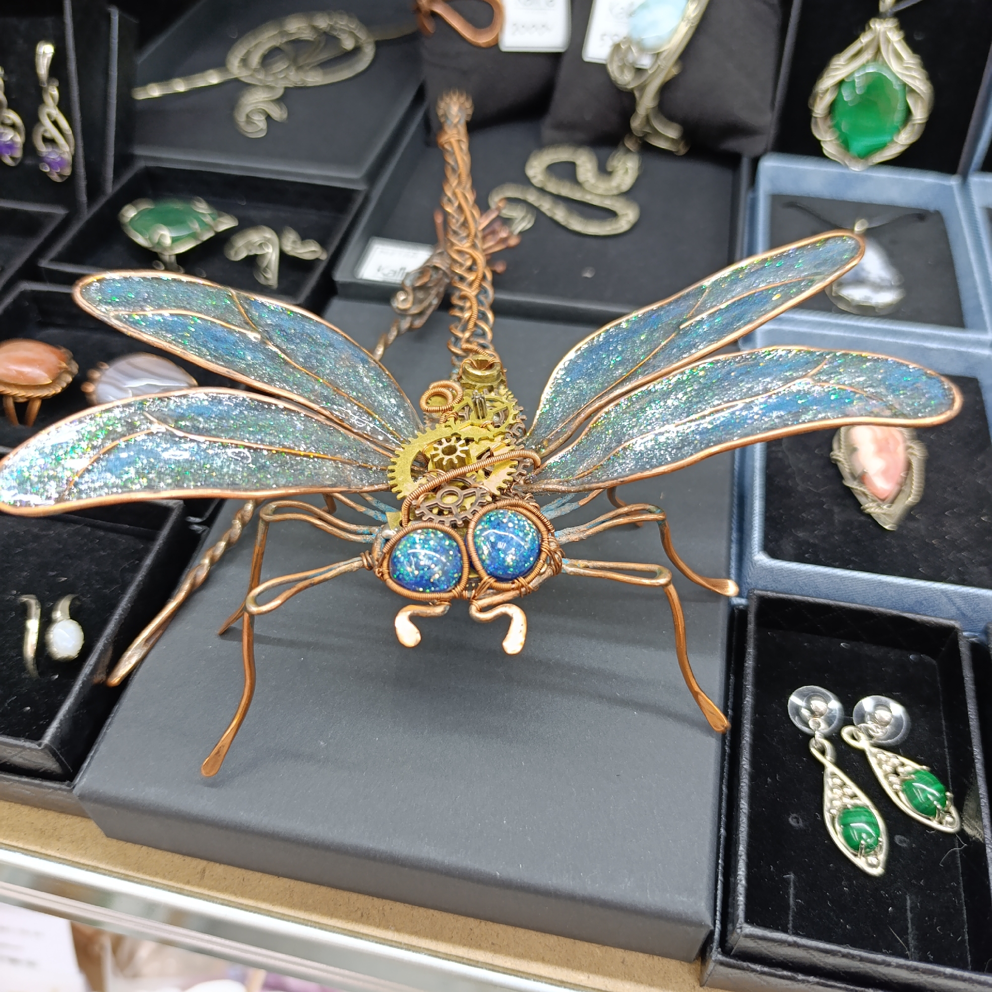 A handcrafted dragonfly ornament made of copper wire, gears, and blue glitter sits in a black jewelry box among other jewelry items in boxes. The dragonfly has wire legs, blue eyes with glitter, gears near its head, and glittery blue wings. The surrounding jewelry includes rings, earrings, and pendants, displayed in black and blue jewelry boxes. The scene appears to be a display in a retail setting, possibly a craft fair or store. The location of Chaikovsky, Russia is not directly indicated but may be implied as the origin or setting for the jewelry.