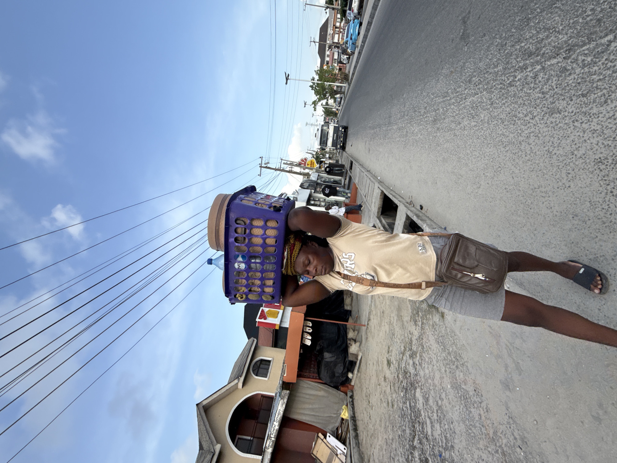 A medium-wide shot displays a woman carrying a basket on her head while walking along a street in Ado, Nigeria. The woman wears a light-colored t-shirt with a dark logo, gray shorts, and black sandals. A brown cross-body bag hangs from her shoulder. Atop her head, she carries a blue plastic basket containing various items, including a clear water bottle. She appears to be walking towards the end of a road. To her left, a building painted with beige, orange, and black is partially visible. A concrete sidewalk borders the road on the right, where the woman is walking. A gray road stretches out ahead of her, lined with cars and buildings in the distance. Overhead, numerous electrical wires crisscross against a backdrop of a blue sky.
