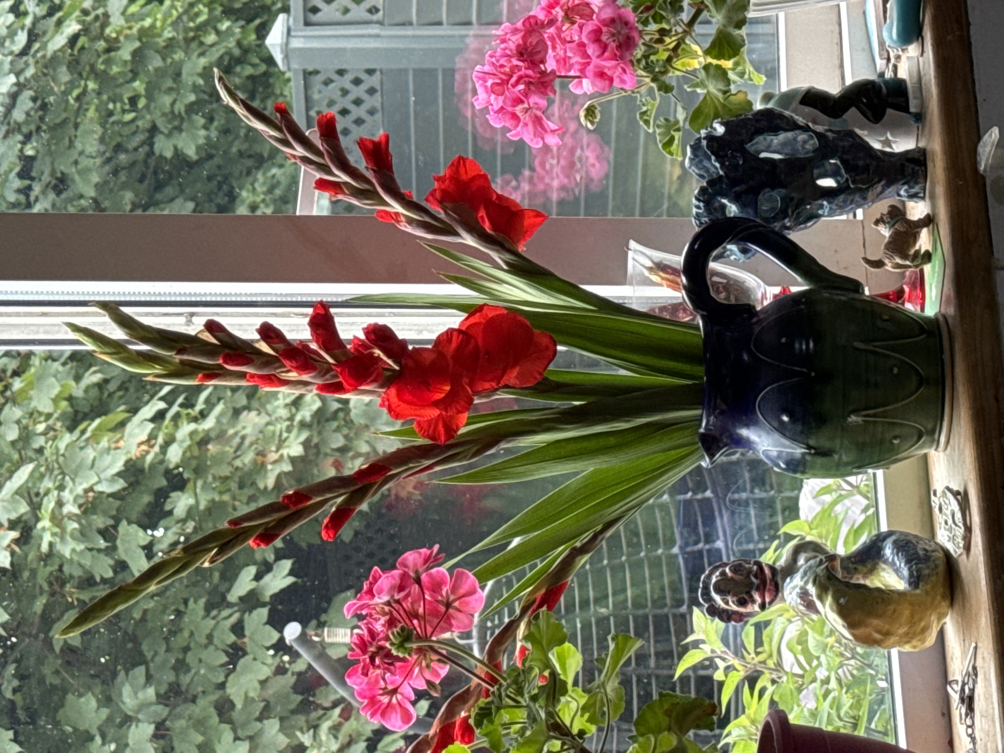 The image displays a still life arrangement on a windowsill, likely in a residential setting, possibly in Waterford, Ireland given the context of the query. In the foreground, a dark blue and green jug holds a bouquet of vibrant red gladioli. To the left of the jug, a ceramic sculpture of a figure with dark hair and facial features, and a yellow and green body, sits on the windowsill. Adjacent to the sculpture is a small metal object, possibly a keyring with several keys. To the right of the jug, a dark blue, abstract sculptural piece stands. Further to the right, a small, tan-colored figurine of a dog with a blue collar is placed on the windowsill. Behind the dog figurine is a white object with a star pattern. Pink geraniums are visible in pots to the left and right of the gladioli, suggesting an indoor or patio environment. The background shows a window looking out onto lush green foliage and a fence, indicating an outdoor view. The overall scene is bathed in natural light, with soft shadows suggesting overcast or diffused lighting conditions.