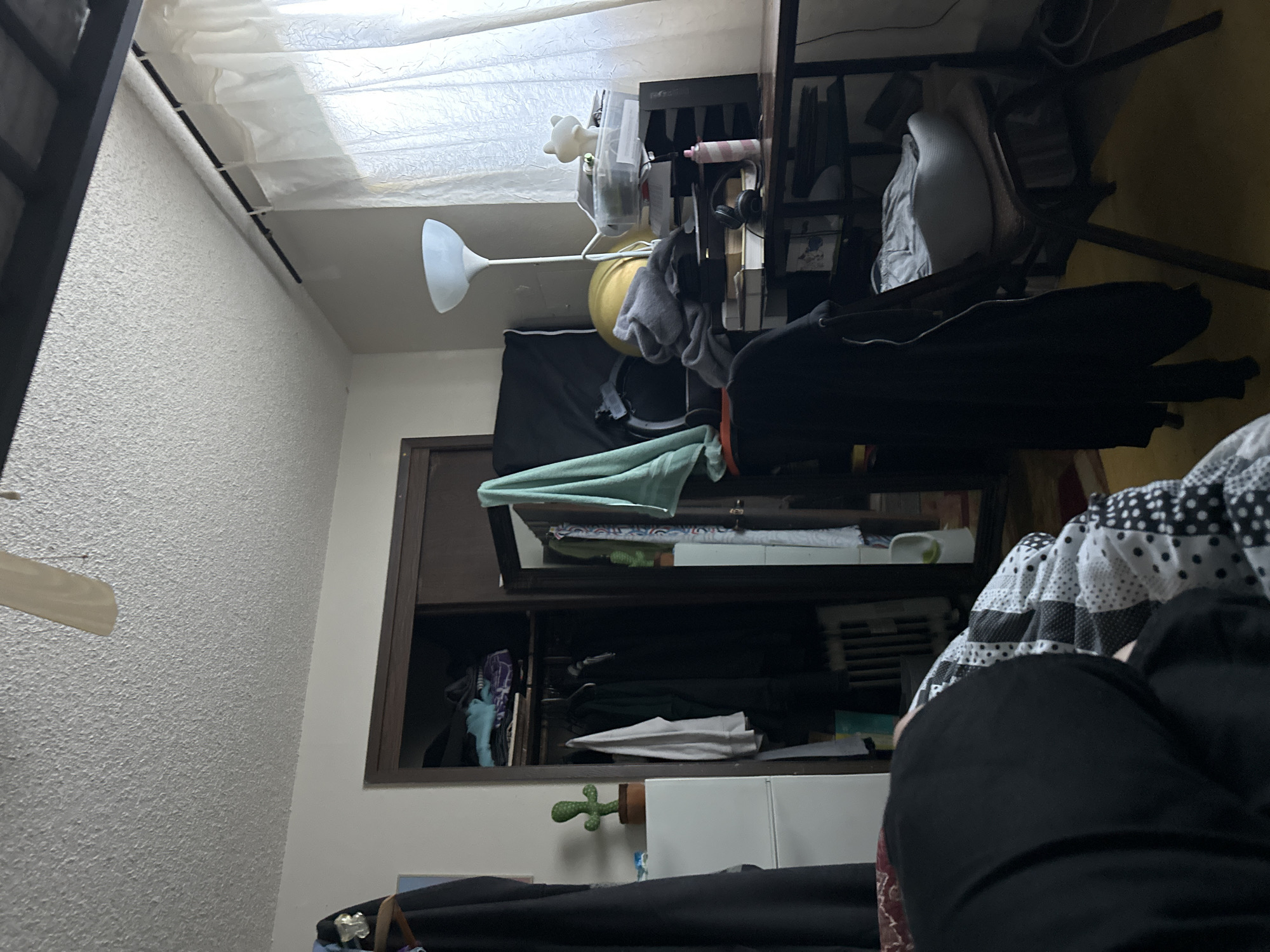 The image depicts a cluttered bedroom. The scene is viewed from a low angle, focusing on a person lying on a bed covered with a black, white, and gray patterned comforter. To the left, a portion of a white dresser is visible with a small potted cactus on top. Adjacent to the dresser is a closet with sliding doors, revealing various clothing items hanging and folded within. On the right side of the image, a desk is covered with numerous items, including papers, containers, headphones, and a pink spray bottle. A floor lamp stands behind the desk, and sheer white curtains hang by a window. The overall impression is one of disarray. The provided context that this scene is located in Vancouver, United States of America is insufficient for a definitive forensic analysis of the location.