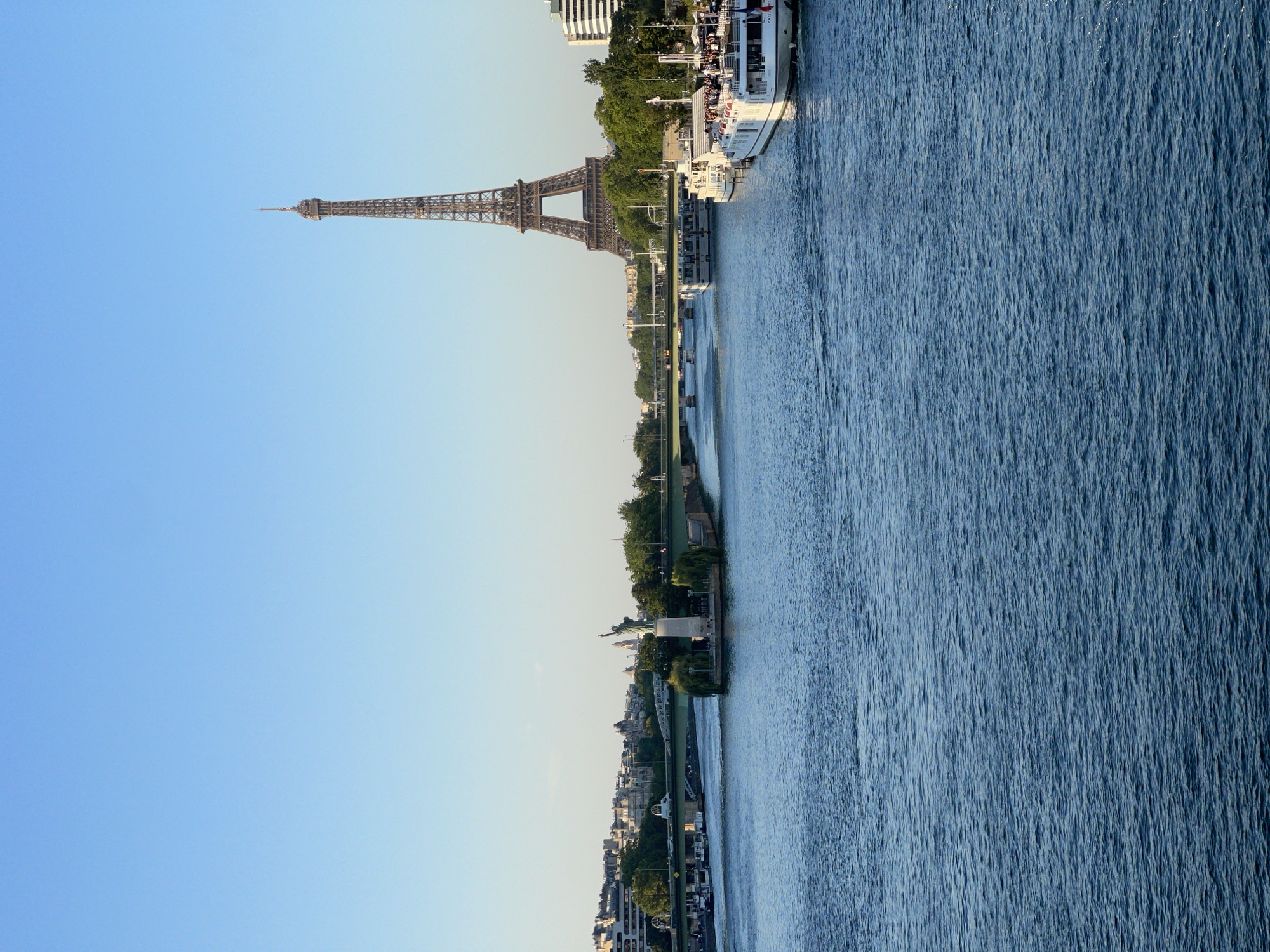 The image captures a panoramic view of the Seine River in Paris, France, with the iconic Eiffel Tower prominently featured in the background. The river occupies the foreground, its surface rippling with a deep blue hue, reflecting the clear sky. Several white passenger boats, some appearing to be tour boats, are moored along the riverbanks or cruising on the water. On the left bank, a bridge spans the river, and beyond it, a city skyline with buildings and trees is visible. In the mid-ground, on a small island in the river, a replica of the Statue of Liberty stands, accompanied by lush greenery. The Eiffel Tower, a lattice-work iron monument, dominates the right side of the background, its intricate structure clearly discernible against the bright, cloudless blue sky. The overall scene depicts a serene day in Paris, with the city's landmarks and waterways bathed in natural light.