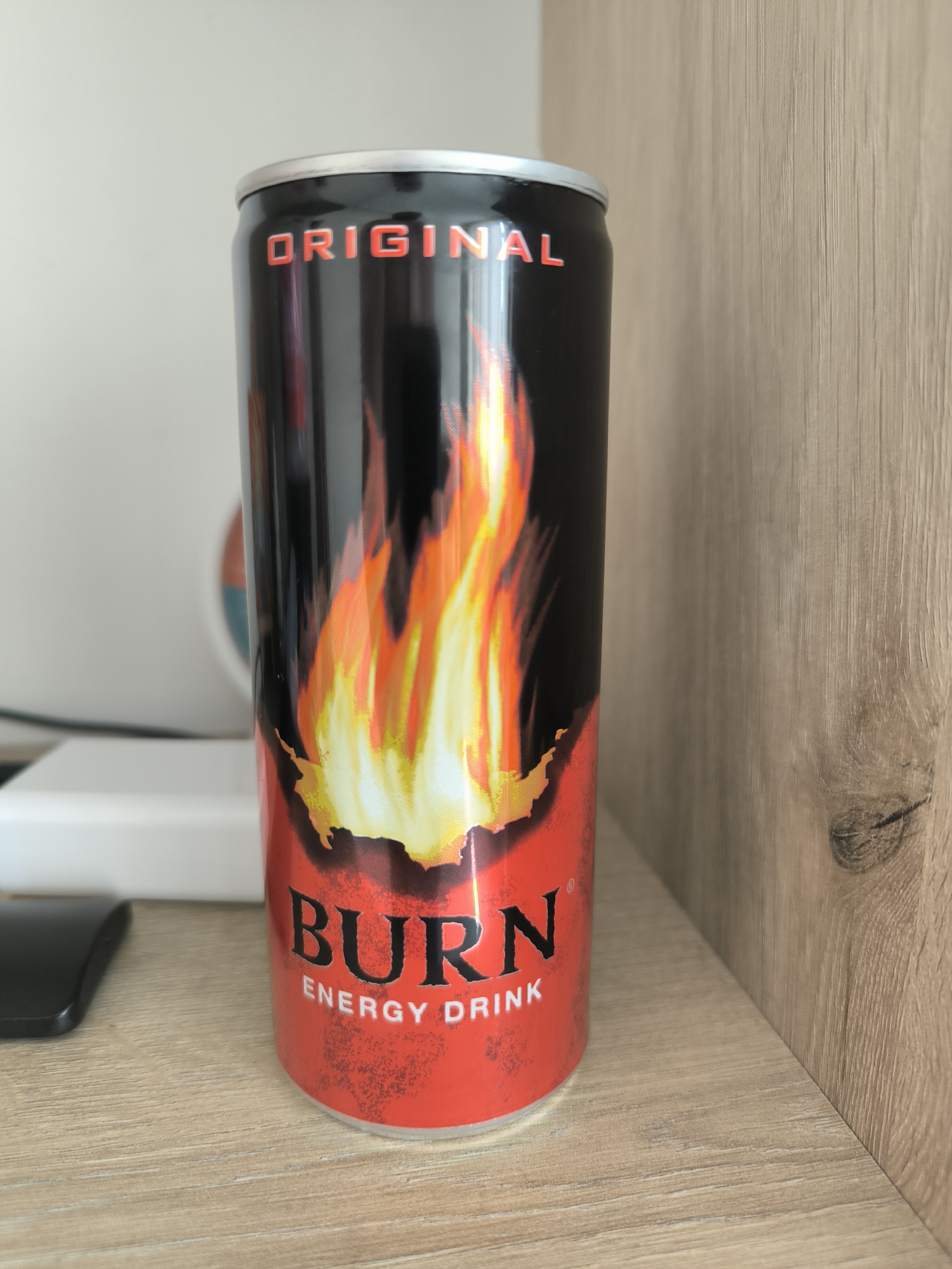 A close-up, eye-level, vertical shot shows a black and red can of "BURN ENERGY DRINK" with a flame design, standing on a light brown, wood-grain textured shelf. The can is positioned slightly off-center to the right. The top of the can features a silver rim. Above the flames, the word "ORIGINAL" is written in red lettering. Below the flames, "BURN" is prominently displayed in large black letters with a red outline, followed by "ENERGY DRINK" in smaller white lettering. The background to the left of the can is blurred, showing a white object that appears to be a router or external hard drive, and a hint of a circular graphic. To the right of the can, a light brown, wood-grain textured wall or vertical surface is visible, with a distinct vertical grain pattern. The lighting is even and appears to be from an overhead source, creating subtle highlights on the can. There are no people or animals present in the image. The scene is indoors, and the overall aesthetic suggests a domestic or office environment. There is no indication of the location being in Chișinău, Moldova.