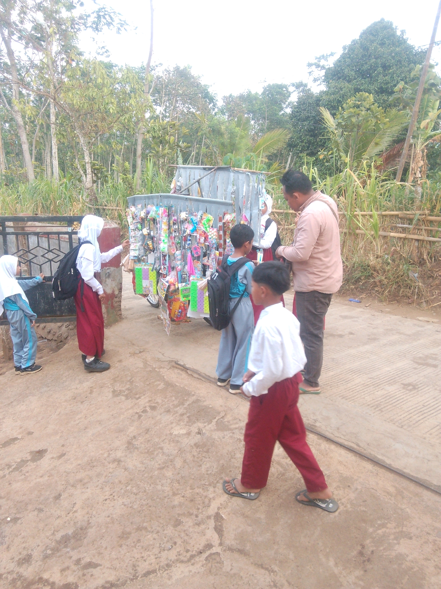 In Betek, Indonesia, children in school uniforms are gathered around a mobile cart laden with merchandise. A young girl wearing a white hijab and maroon pants stands to the left, reaching towards the cart. Beside her, another child in a grey uniform looks on. In the background, a man in a light pink shirt and dark pants stands near the cart, appearing to interact with the children. A boy in a white shirt and maroon pants walks away from the cart towards the foreground, while another boy in a grey uniform with a backpack stands to the left of the man. The cart is stocked with various colorful items, likely snacks or small toys. The scene takes place outdoors on a paved surface, with a backdrop of lush green vegetation and trees.