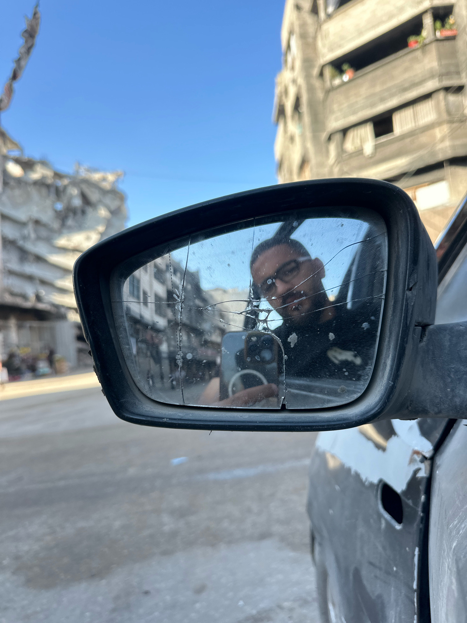 A cracked side-view mirror of a vehicle reflects a man wearing glasses and holding a mobile phone. The mirror is mounted on the exterior of a dark-colored car, which appears weathered and possibly damaged. In the background, out of focus, are damaged buildings characteristic of war-torn urban environments. To the left, a partially destroyed structure with debris suggests significant damage. To the right, a taller, concrete building with balconies and windows is visible. The sky is clear and blue. The overall scene appears to be set in the city of Gaza, Palestinian Territories, given the context of destroyed buildings and the likely location associated with such imagery.