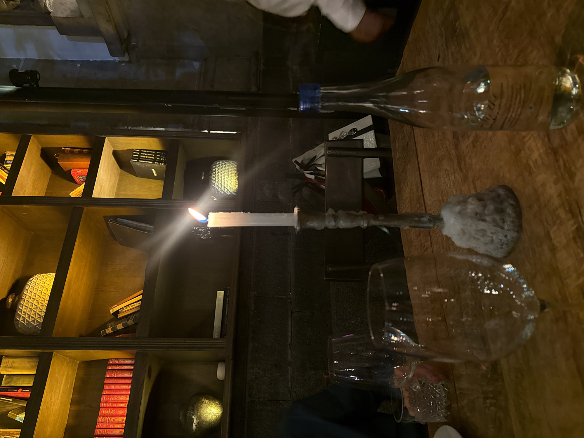 The image captures a scene in what appears to be a dimly lit restaurant or bar. A table with a wooden surface occupies the foreground, on which there is a water bottle with a blue cap, a lit candle in a silver candlestick, and a partially visible wine glass. The lighting from the candle creates a strong contrast with the rest of the scene. In the background, a bookshelf filled with books and decorative objects provides an interesting backdrop. Based on the style and ambiance, as well as a previous query, the location is likely in Mexico City, Mexico. The image suggests a setting of a restaurant or bar with a unique ambiance.
