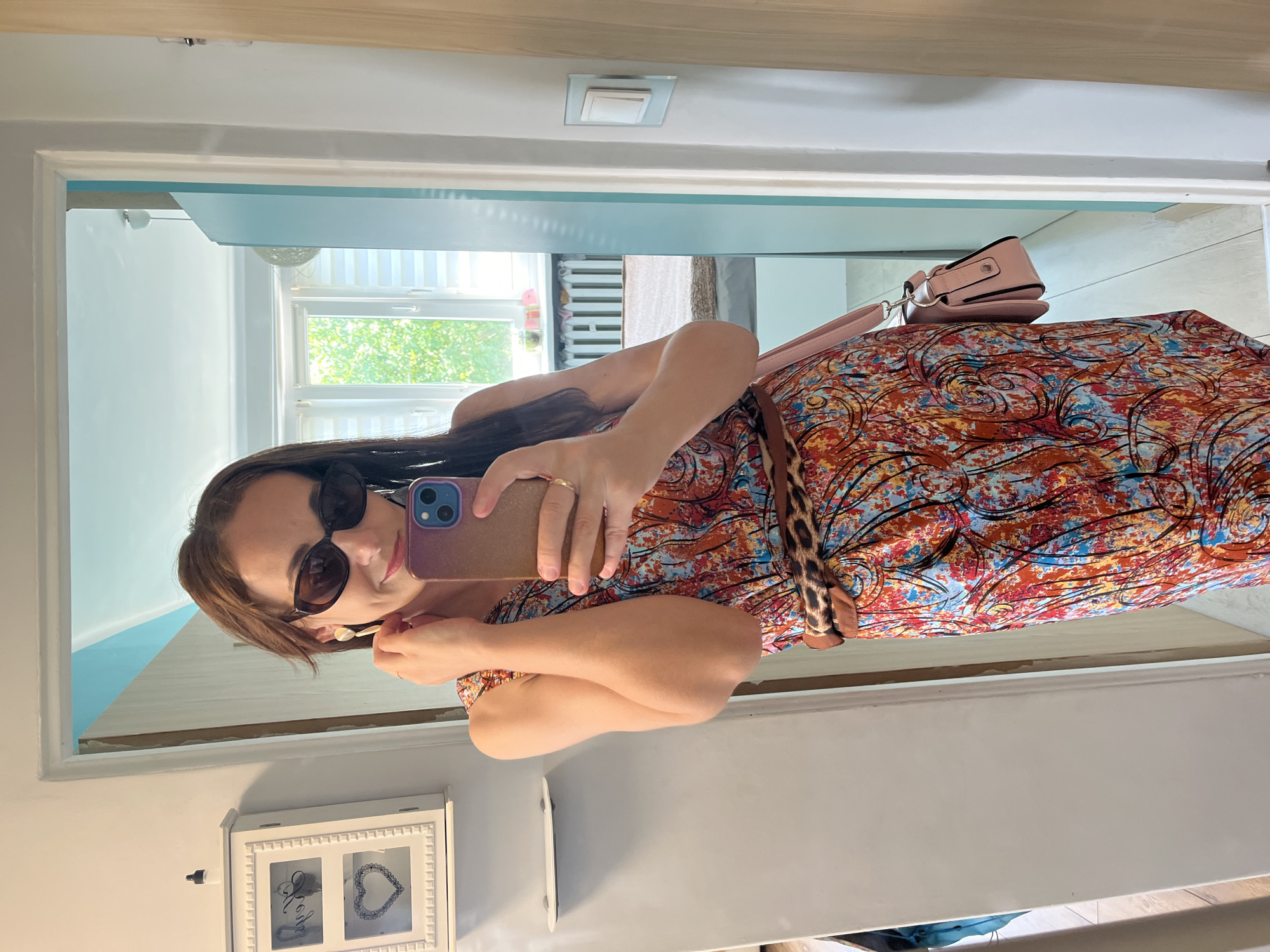 A woman with brown hair, wearing sunglasses and a colorful, patterned dress, takes a selfie in a mirror. She is holding a smartphone in her right hand, with her left hand resting on her hip. A pink purse is slung over her shoulder. A leopard-print belt cinches her waist. The background of the mirror shows a room with a window, blinds, and a decorative frame with hearts. The lighting suggests it is daytime, possibly indoors. The woman appears to be posing for the photo. There is no visible text in the image.