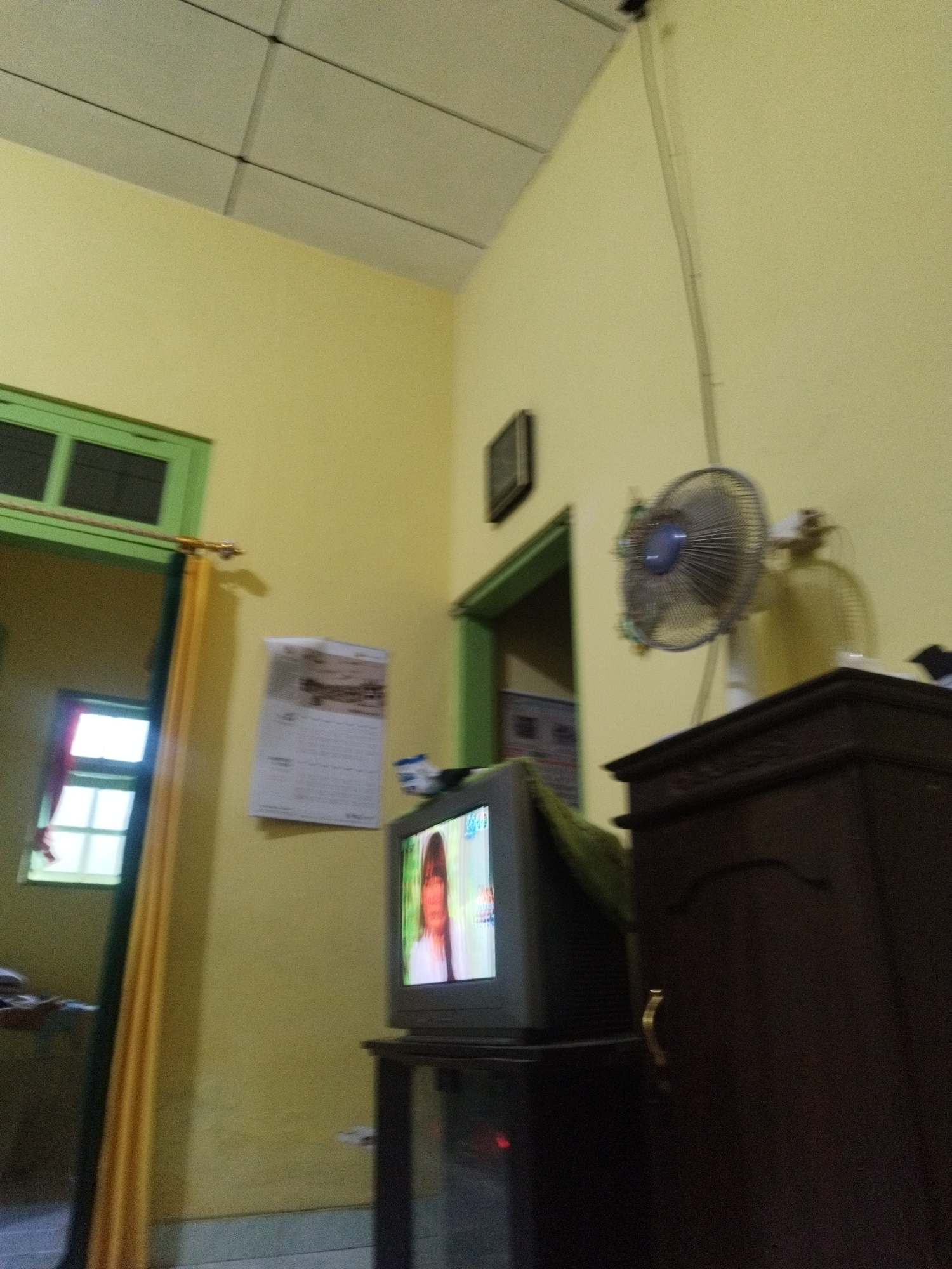 The image depicts an indoor scene in a room with pale yellow walls and a white tiled ceiling. A vintage television set is placed on a dark wooden stand, displaying a person's face. To the right of the television, a dark brown wooden cabinet with a decorative brass handle is visible. On the wall above the television, a calendar is hanging, and a ceiling fan is mounted. A doorway with a green frame is seen in the background, leading to another room. A window with a green frame is also present, showing a glimpse of the outside. The overall impression is that of a simple, lived-in domestic space. The specific location in Sumenep, Indonesia cannot be determined from the visual information alone.