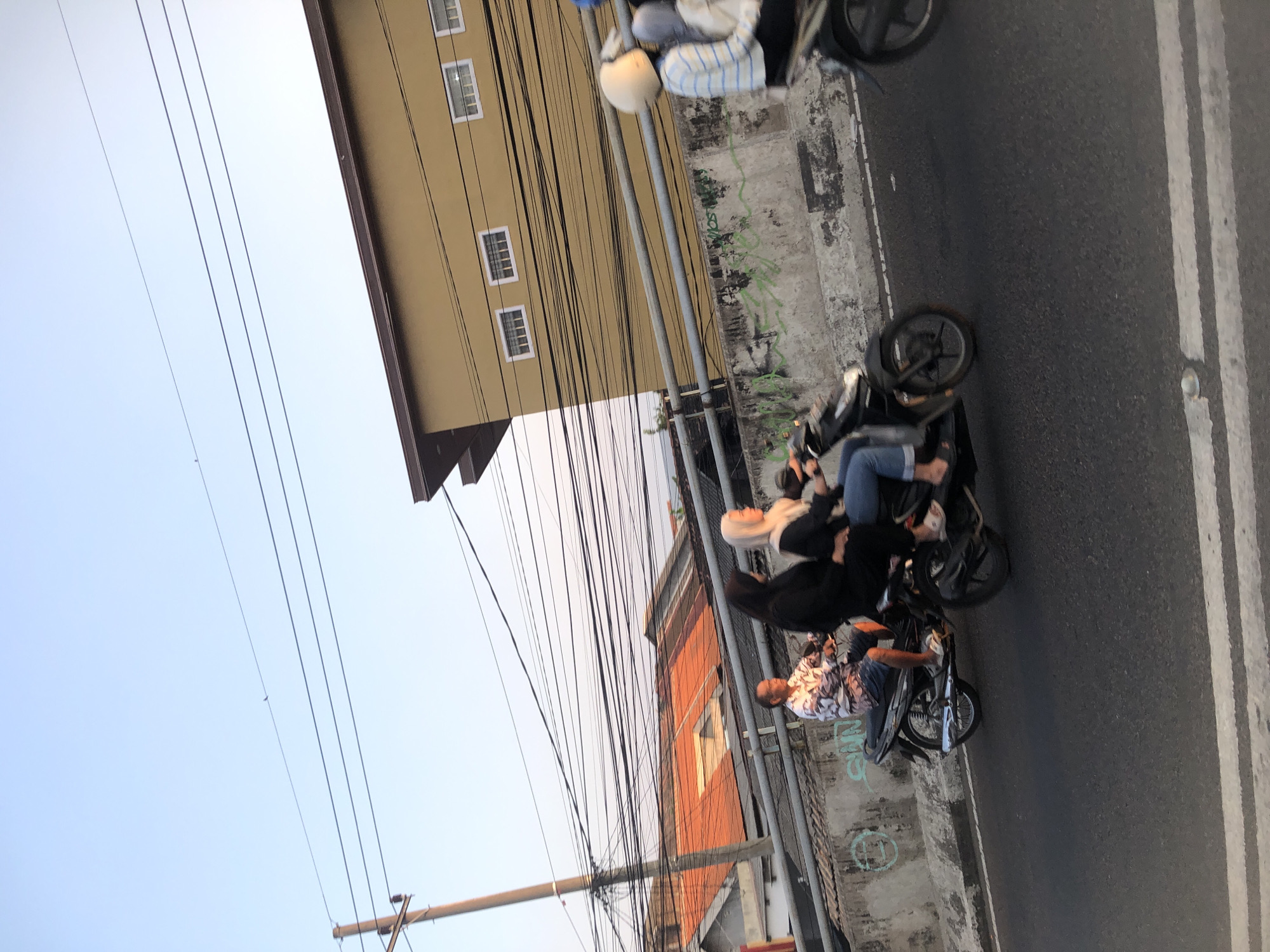 A medium shot captures a street scene in Tenggulunan, Indonesia, with the sun casting a warm glow. In the foreground, two motorcycles are in motion on an asphalt road, indicated by motion blur. The first motorcycle, on the left, carries a man wearing a patterned shirt and shorts, and a woman in a black hijab. The second motorcycle, to the right, has two individuals. The rider is partially visible with a white helmet and a striped long-sleeved shirt. The passenger is wearing a light-colored hijab and jeans. Behind the motorcycles, a concrete barrier with graffiti runs along the side of the road. In the background, an orange-roofed building is visible, alongside a yellow, multi-story building with several windows. A complex network of electrical wires crisscrosses the sky, supported by a wooden utility pole on the left. The sky is clear and blue, with a hint of a sunset's warmth.