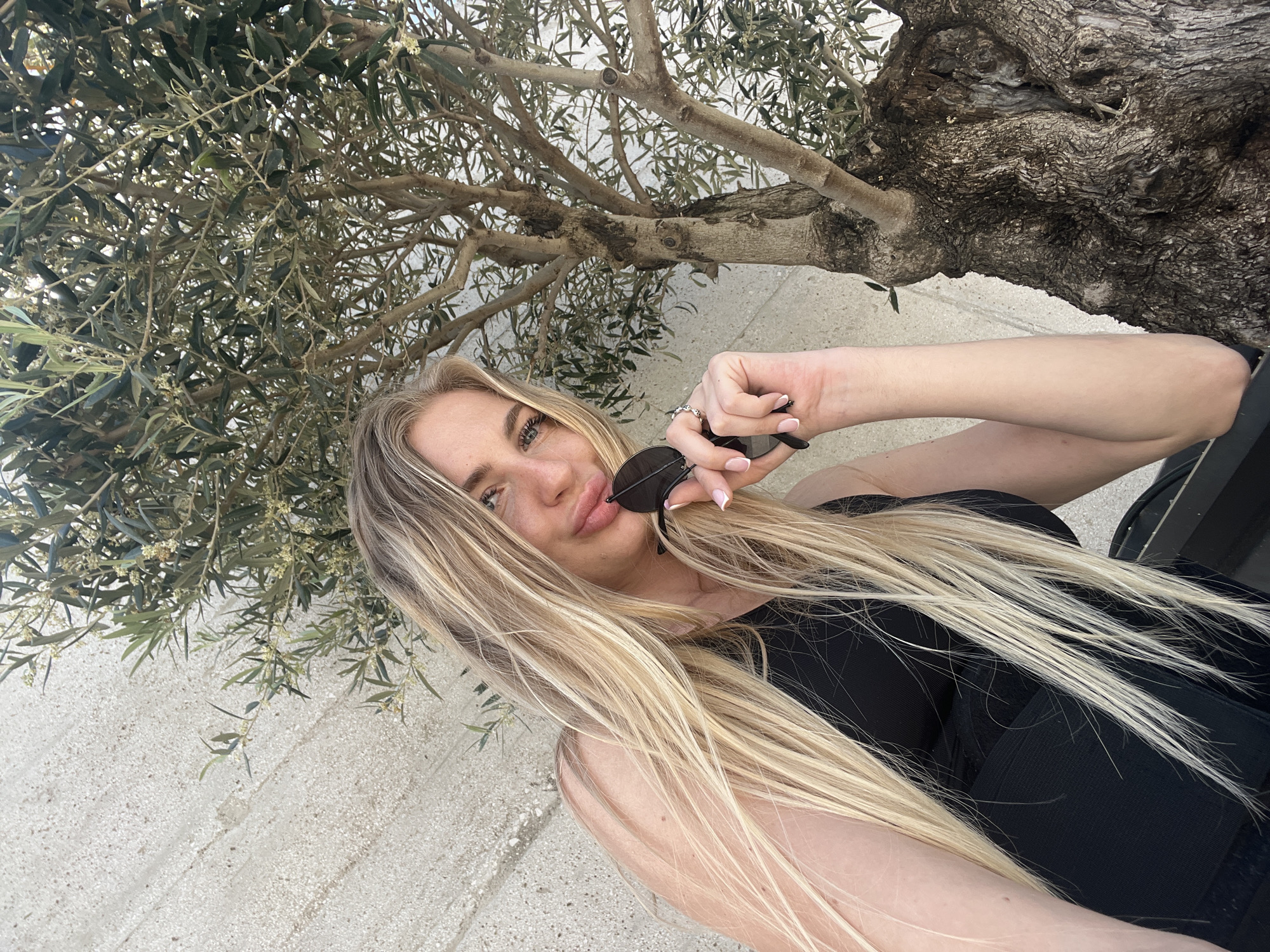 A woman with long, blonde hair poses for a selfie outdoors. She is wearing a black top and holding sunglasses in her hand. In the background, there is a tree with textured bark and green leaves. The ground appears to be a light-colored stone or concrete. The image was likely taken in Radovići, Montenegro.