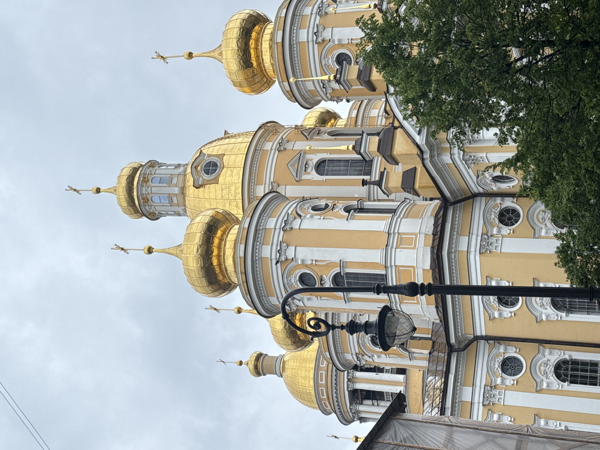 The image shows a fragment of the Saint Nicholas Naval Cathedral, a major Russian Orthodox cathedral in Saint Petersburg, Russia. The cathedral is a baroque structure with prominent golden domes and yellow-and-white facades. A black lamppost is situated in the foreground, obscuring a portion of the building. A tree with green leaves is visible to the right of the frame. The sky is overcast, suggesting a cloudy day. The orientation of the image is vertical.