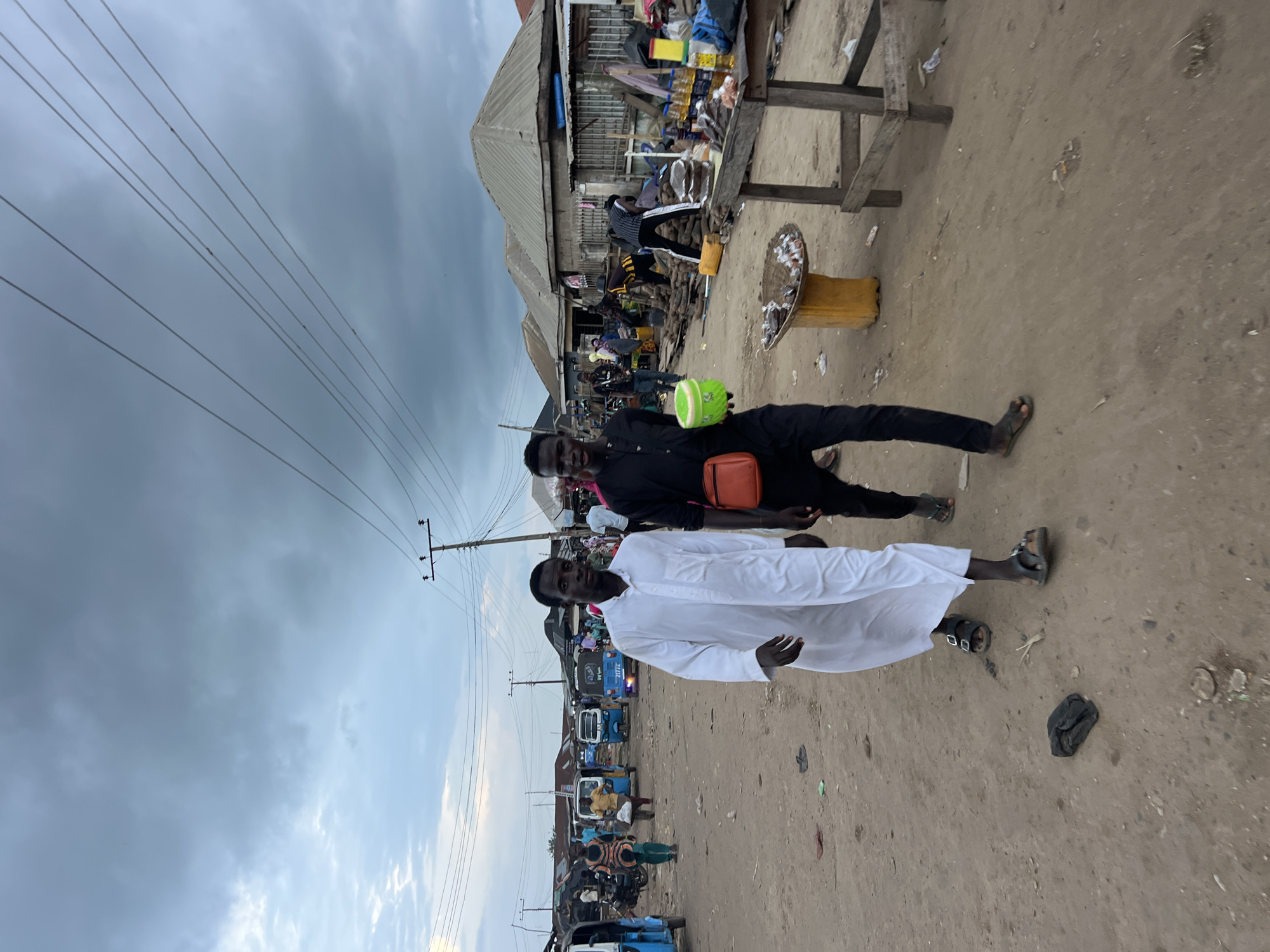 This is a medium shot, eye-level, outdoor photograph taken in the city of Oko Amakom, Nigeria. The scene depicts a dirt road lined with market stalls and buildings. The sky is overcast with heavy clouds. Two young men are walking towards the camera in the foreground. The man on the left is wearing a long white robe and sandals. The man on the right is wearing a black long-sleeved shirt and pants, also with sandals, and is carrying a bright green bucket and an orange bag across his chest. In the background, several blue and yellow motorized tricycles, known as "keke," are parked or moving. There are various market goods displayed on wooden tables and on the ground, including produce like yams, bottles of oil, and other items. Several other individuals are visible in the background, some appearing to be vendors and customers interacting in the marketplace. The overall impression is of a busy, local market day.