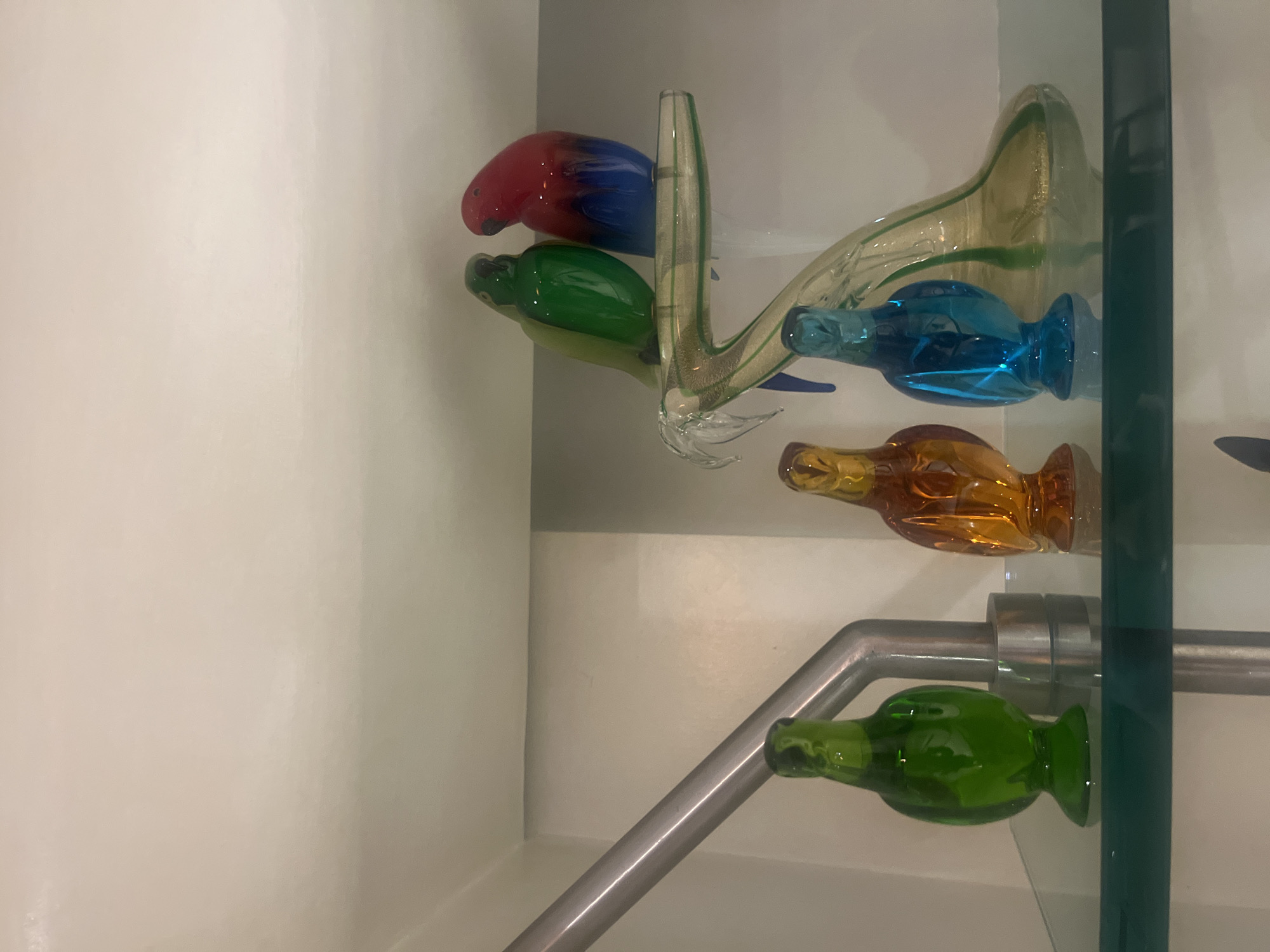 The scene depicts an indoor display of five glass bird figurines. On a clear glass branch sculpture, positioned centrally on an upper transparent shelf, a green glass parrot faces a bicolored red-to-blue glass parrot. Below and to the left on the same transparent shelf, a standalone green glass bird figurine is present. To its right, an amber-colored glass bird figurine is positioned next to a blue glass bird figurine. A silver-colored metal pipe, bent at an angle, is visible on the left side, extending upwards behind the leftmost bottom figurine. A horizontal green line runs across the lower portion of the frame, indicating a shelf or structural element below the main display. The background consists of plain, light-toned walls. The environment is uniformly and brightly lit. No specific identifying details within the image indicate a precise location within Mexico City, Mexico.