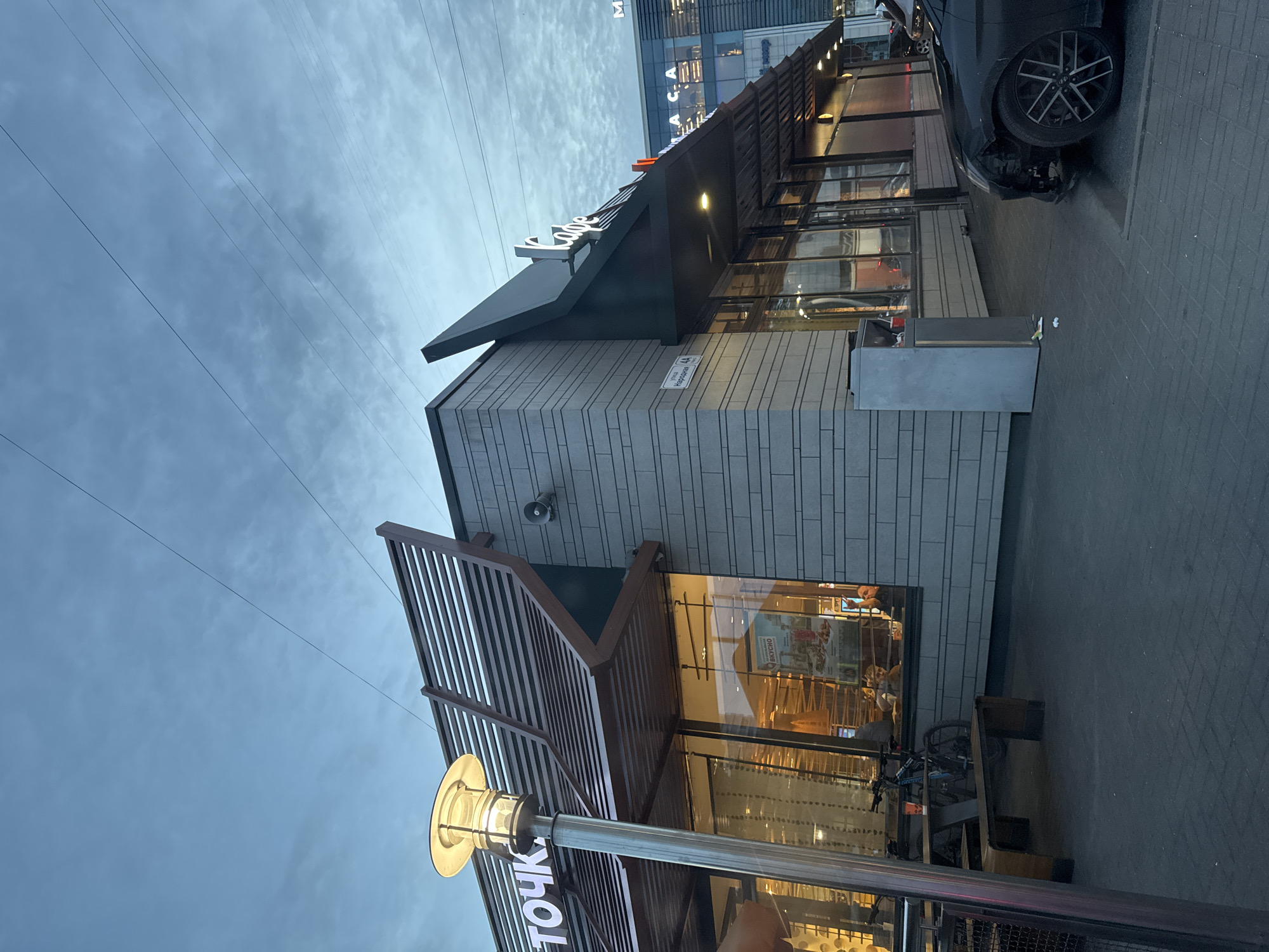 A commercial establishment with signage reading "ТОЧКА." and "Кафе" is visible under a cloudy sky. The building features a modern architectural design with a grey brick facade and a slatted wooden overhang. A street lamp stands to the left of the building, illuminated. Inside the establishment, through large windows, patrons can be seen sitting at tables. In front of the building, a bicycle is parked next to a wooden bench. A black car with damage to its front bumper is parked on the right side of the image, partially obscuring the building's entrance. The ground is paved with grey cobblestones. A sign on the building indicates "ул. Народная, 44," suggesting the location is Narodnaya Street, 44, in Saint Petersburg, Russia. The overall scene depicts an exterior view of a public establishment during twilight.