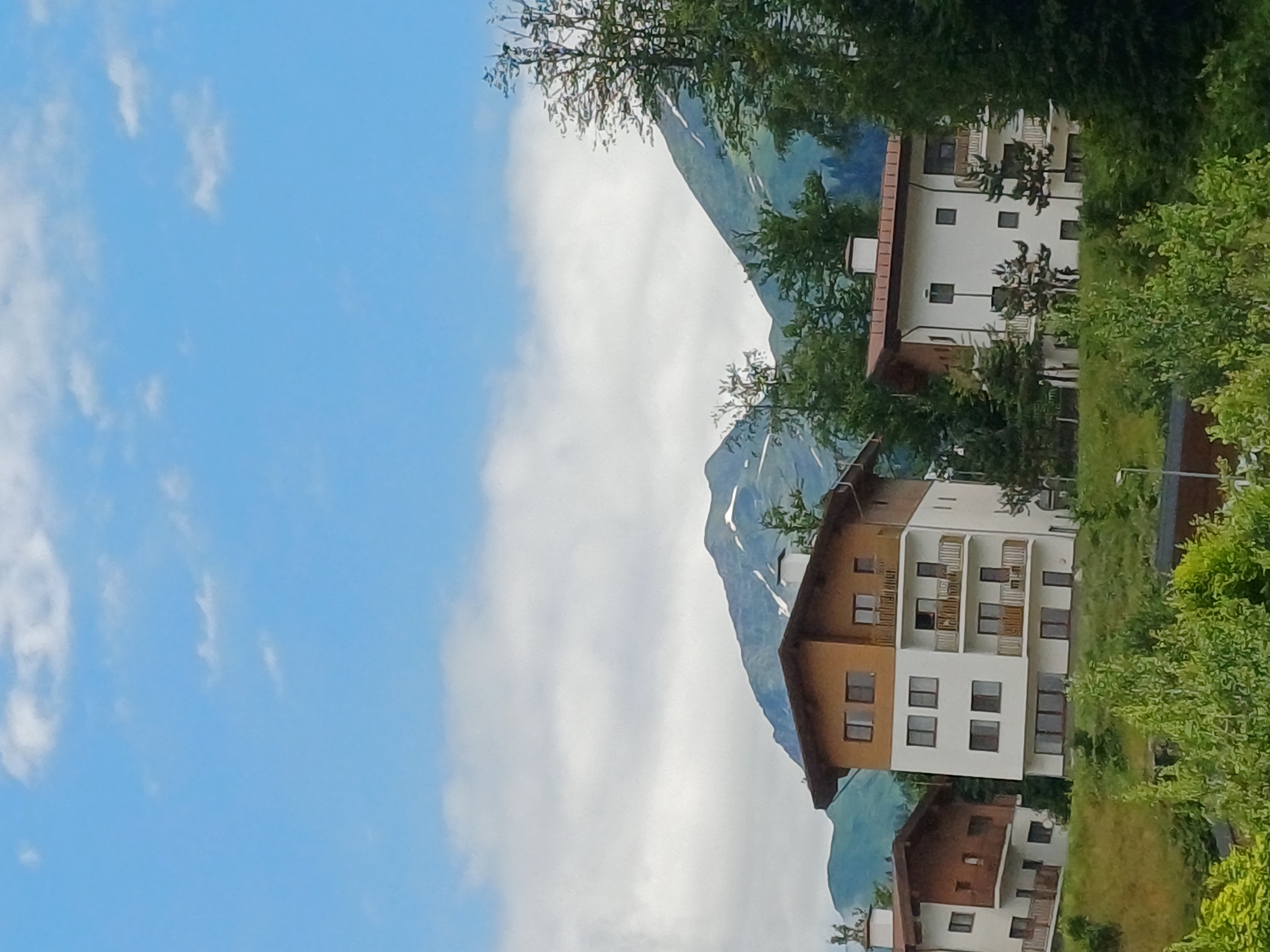 The image depicts a serene mountain landscape with modern, multi-story buildings nestled on a hillside. The sky is a bright blue with scattered white clouds. In the foreground, lush green trees and foliage cover the rolling hills. The buildings are contemporary in design, featuring white facades, brown roofs, and balconies. The architecture suggests a resort or residential area in a mountainous region. The background is dominated by imposing mountains, some of which are capped with snow, indicating a high-altitude location. The scene conveys a sense of peacefulness and natural beauty. This specific scene is located in the city of Estosadok, Russia, known for its ski resorts and picturesque mountain views. There are no people or animals visible in the image, and no specific actions or interactions are depicted. The overall environment is that of a well-developed natural area with tourist or residential infrastructure.