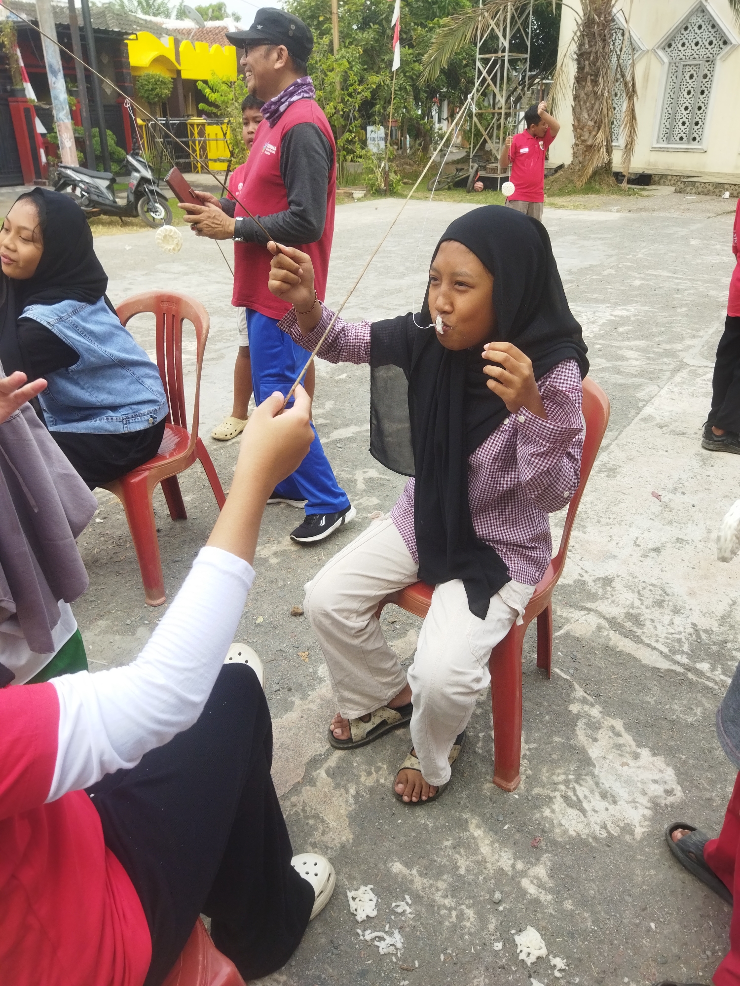 In Banjar, Indonesia, a group of people are engaged in a game. A young girl wearing a black hijab and a checkered shirt sits on a red plastic chair, attempting to pick up a white object with a stick held in her mouth. The object is attached to a string, which is being held by a man in a red shirt and a black cap. Another young girl, also wearing a black hijab and a denim vest, is seated to the left, partially visible. In the background, a man in a red shirt and blue pants stands near a building with decorative windows. The ground is paved with concrete, and debris is scattered around. The environment suggests an outdoor gathering or event.