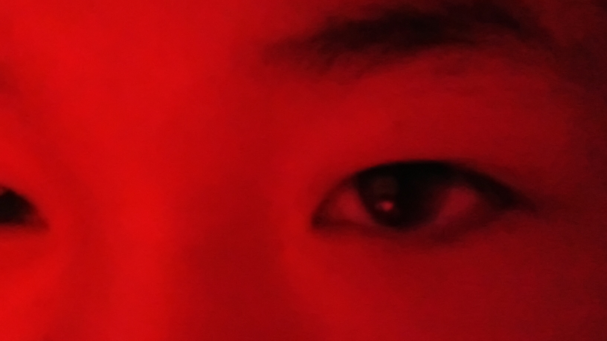 The image is a close-up, abstract photograph of a person's eyes, dominated by a vibrant red hue. The lighting is such that details are obscured, creating a dreamlike or intense atmosphere. The left eye is partially visible on the left side of the frame, while the right eye is more centrally located. The sclera and iris of the right eye are discernible, with a glint of light reflecting in the pupil. The surrounding skin texture is smoothed and indistinct due to the lighting and focus. There is no indication of any urban environment, city, or specific location such as Uchkun, Kyrgyzstan. The image solely focuses on the facial feature of the eyes.